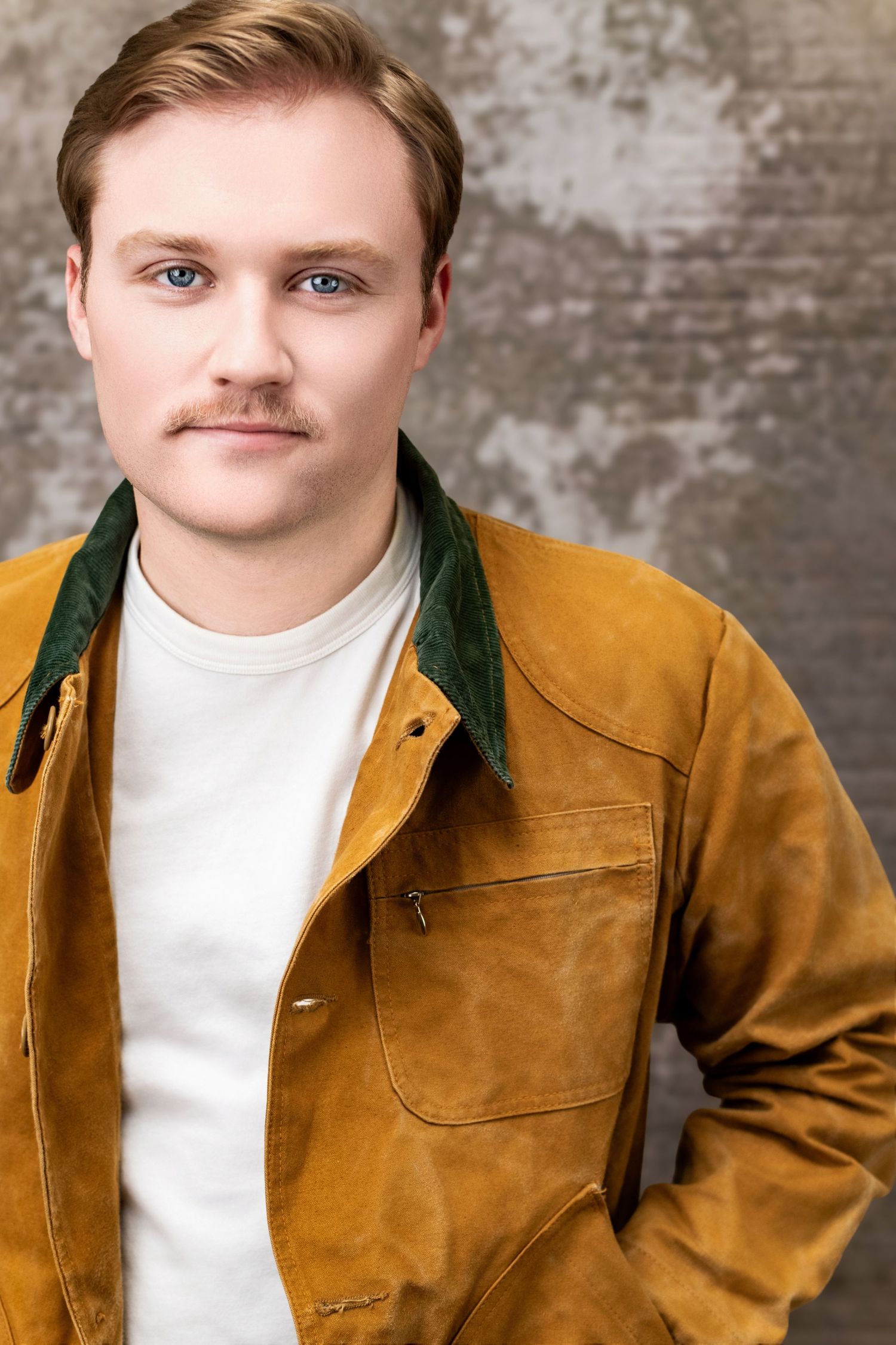 Theatrical headshot of a man with serious expression in mustard jacket, styled for grounded, character-driven roles