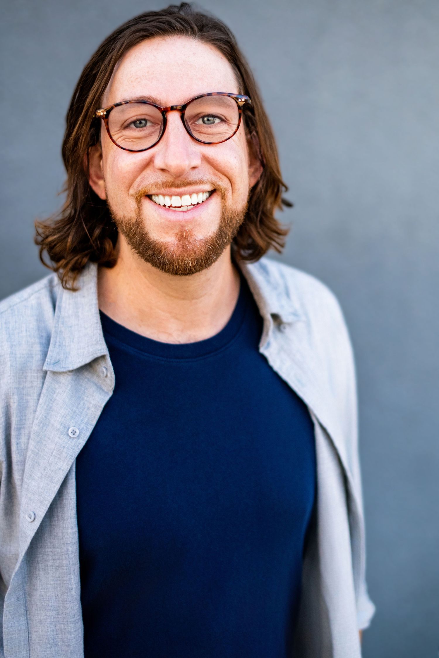 Male business professional wearing glasses and a casual button-down, photographed against a gray backdrop for a professional headshot.