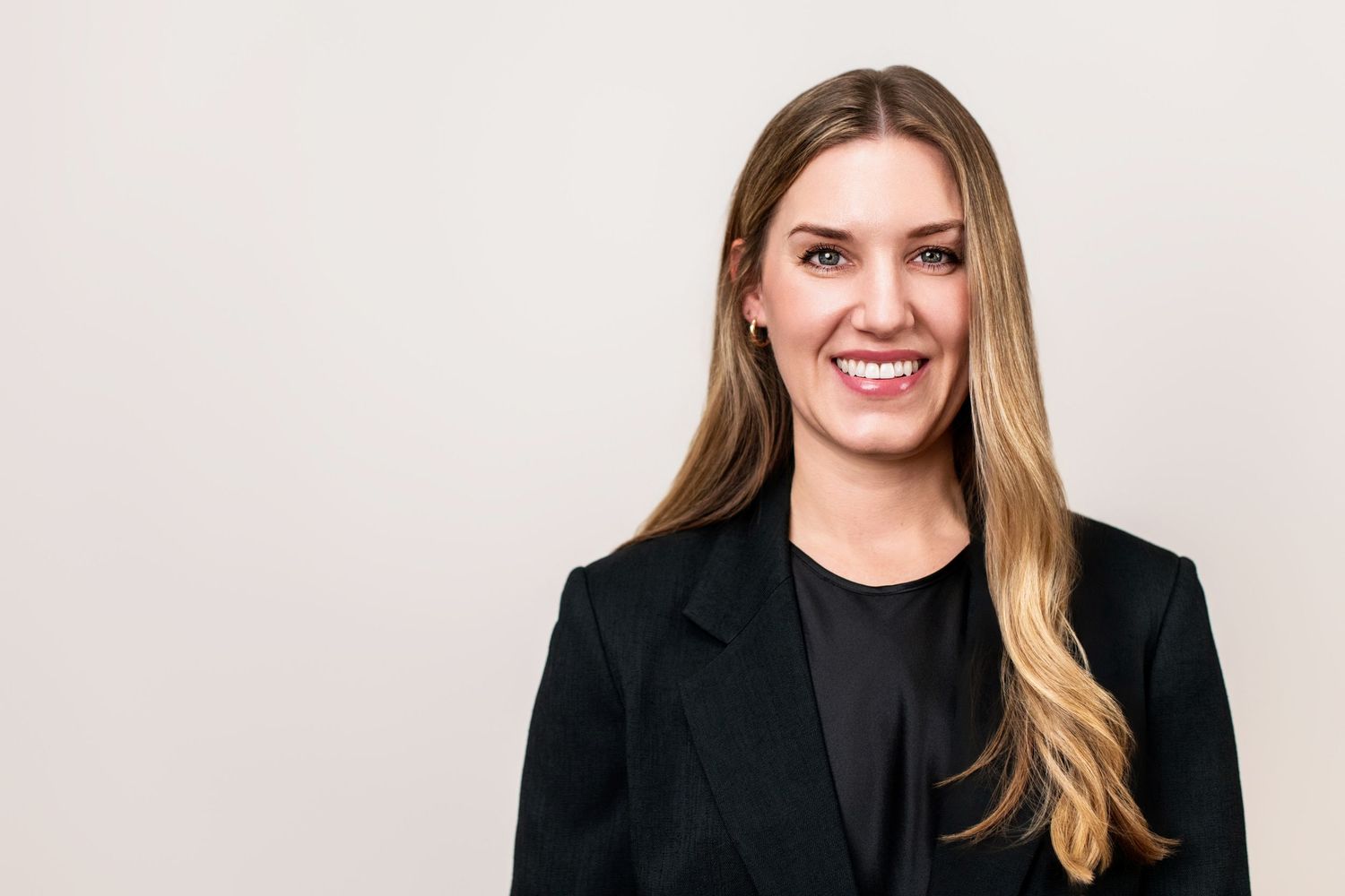 Professional executive headshot of a woman in a black blazer, smiling confidently against a clean neutral studio backdrop.