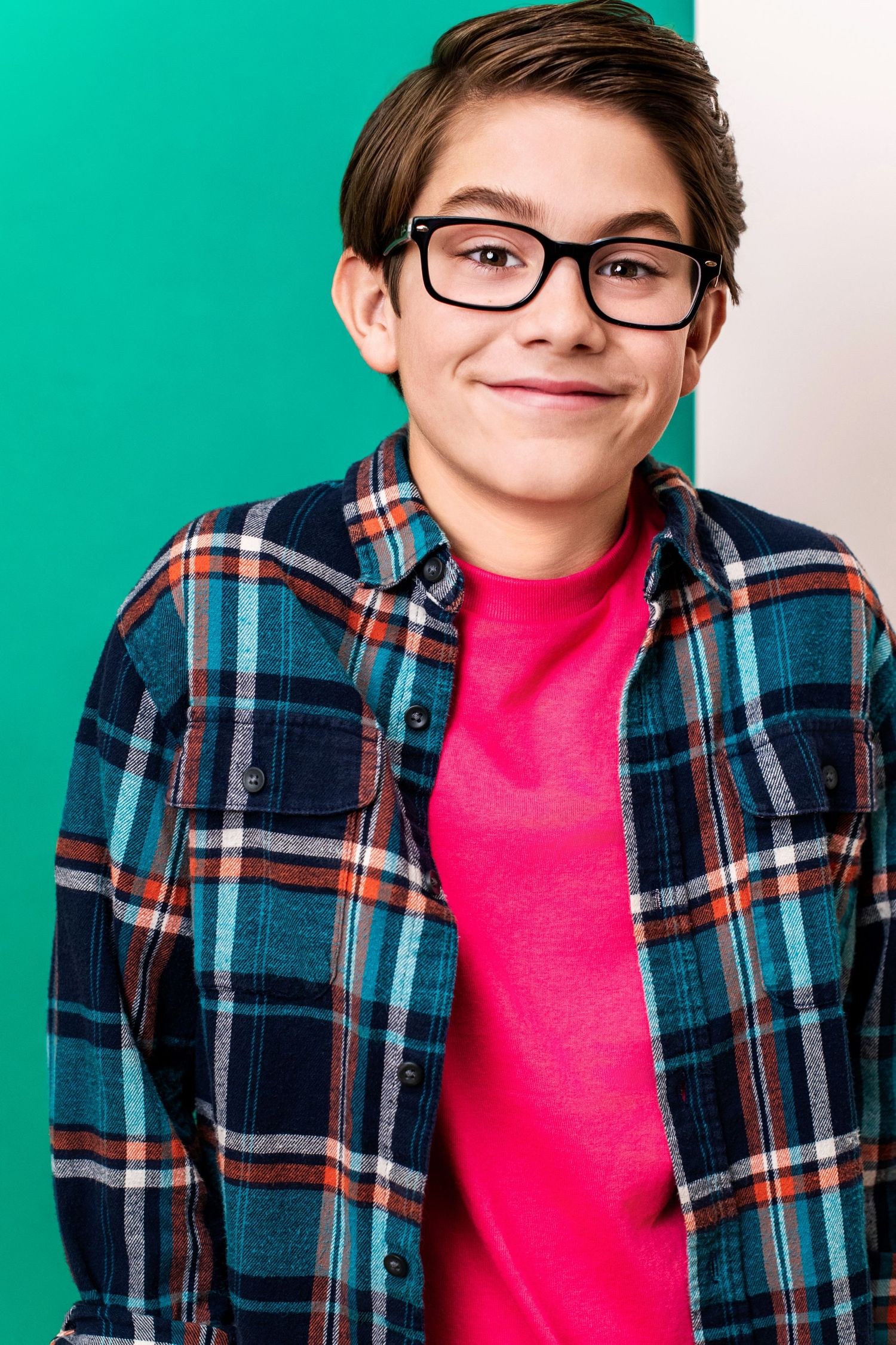 Smiling youth boy wearing glasses and a plaid shirt against a teal background for a professional commercial headshot.