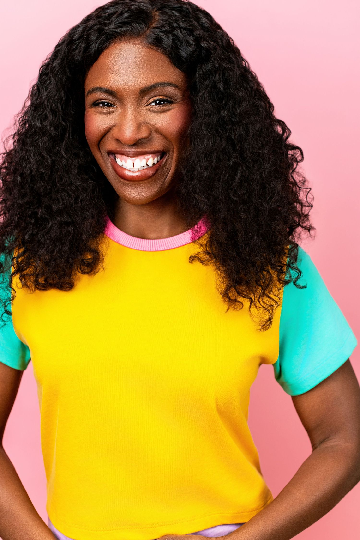 Joyful woman with curly hair wearing a yellow top against a pink background photographed by Nice Headshots.