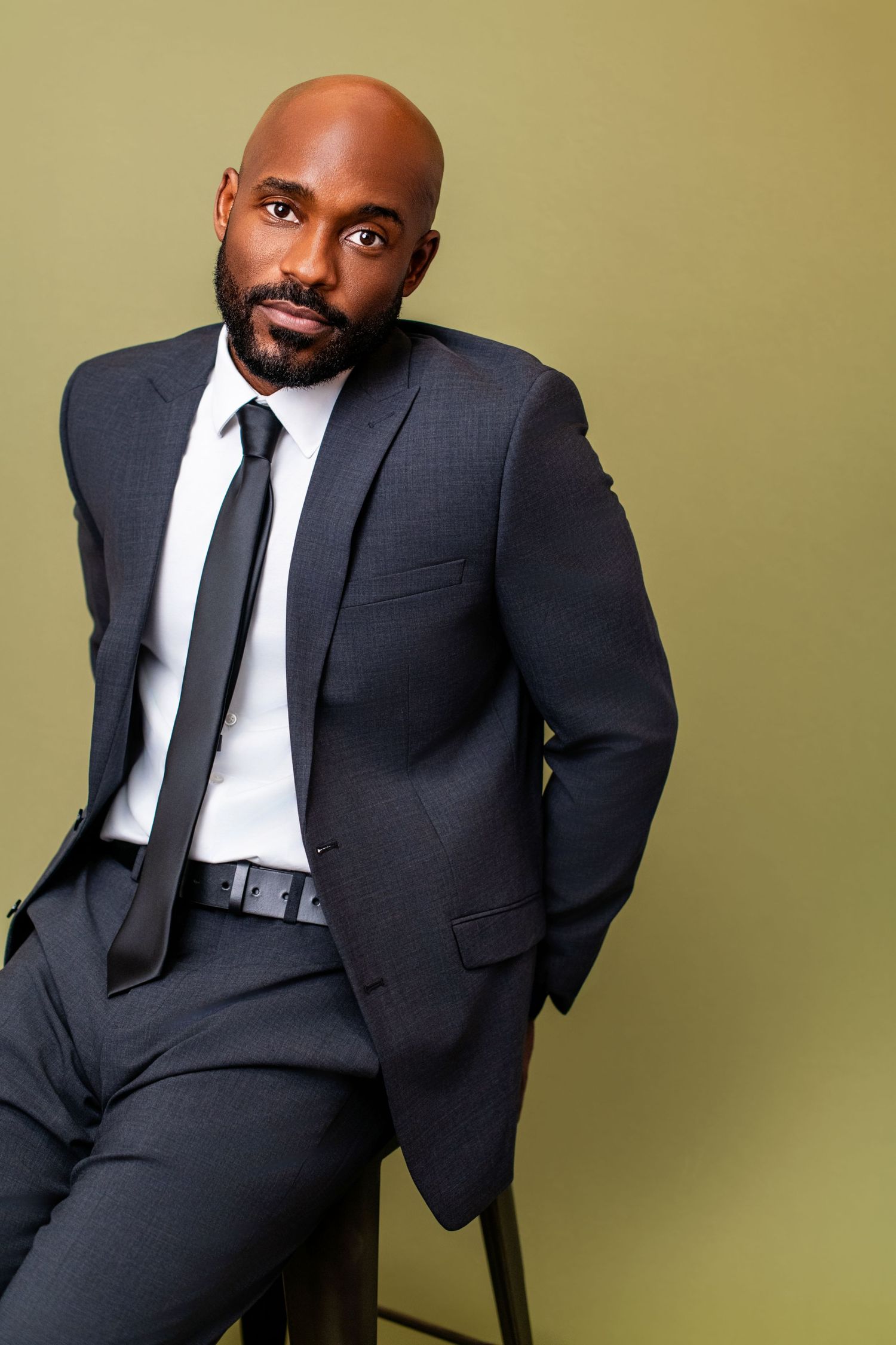 Male model in a suit leaning casually on a stool, photographed against an olive green backdrop for a modeling headshot in Los Angeles