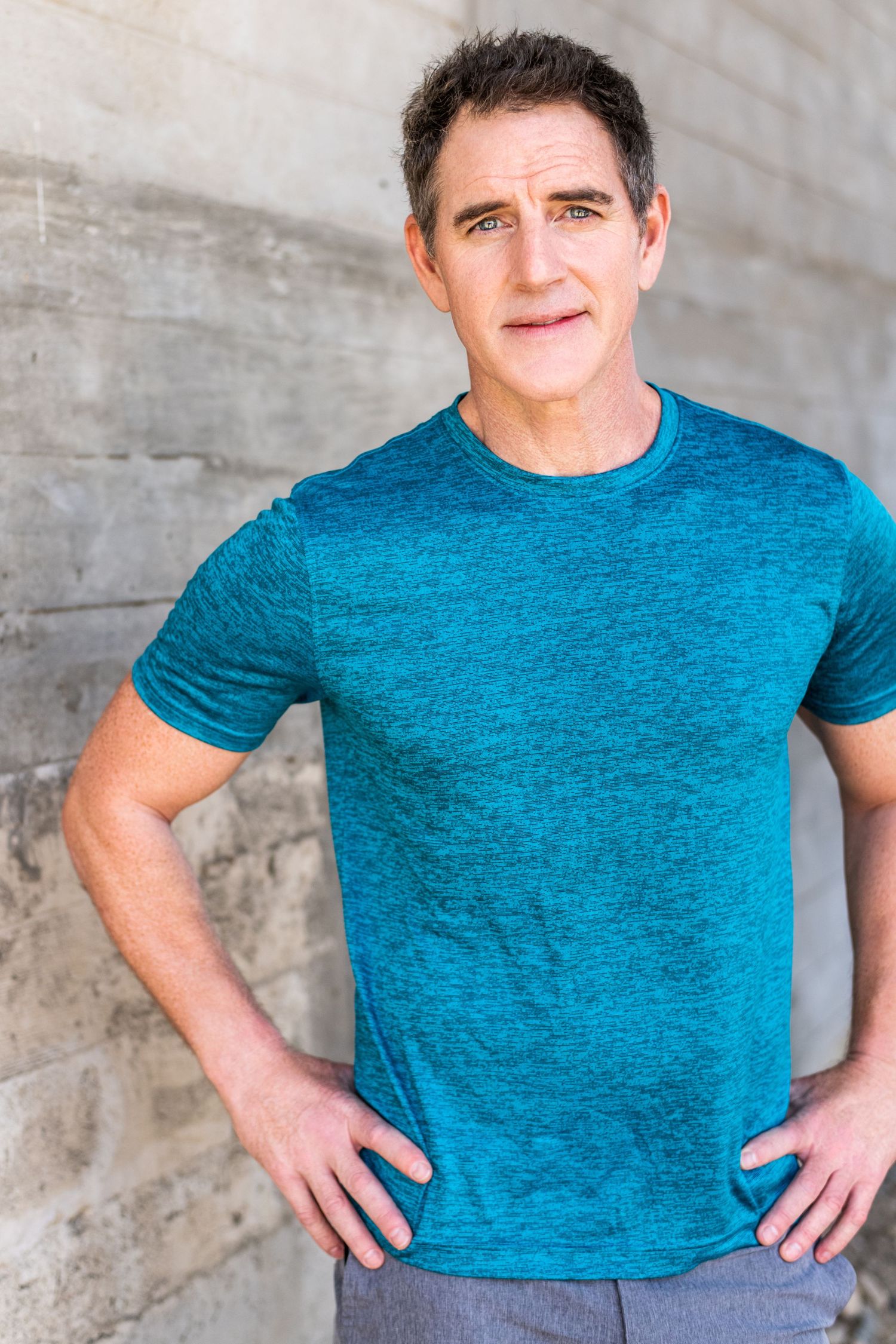 Male model wearing a teal t-shirt with hands on hips, photographed against a concrete wall for a professional modeling headshot in L.A.