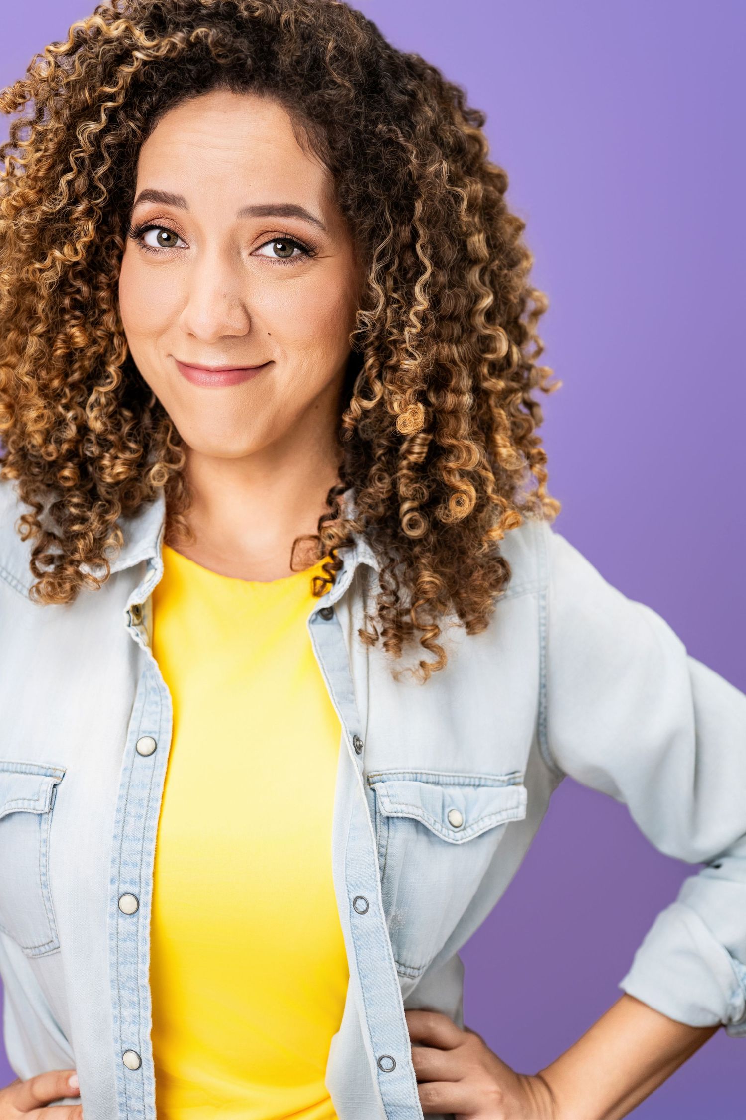 Smiling woman with curly hair wearing a yellow top and denim shirt on a purple background, photographed in a Los Angeles headshot studio