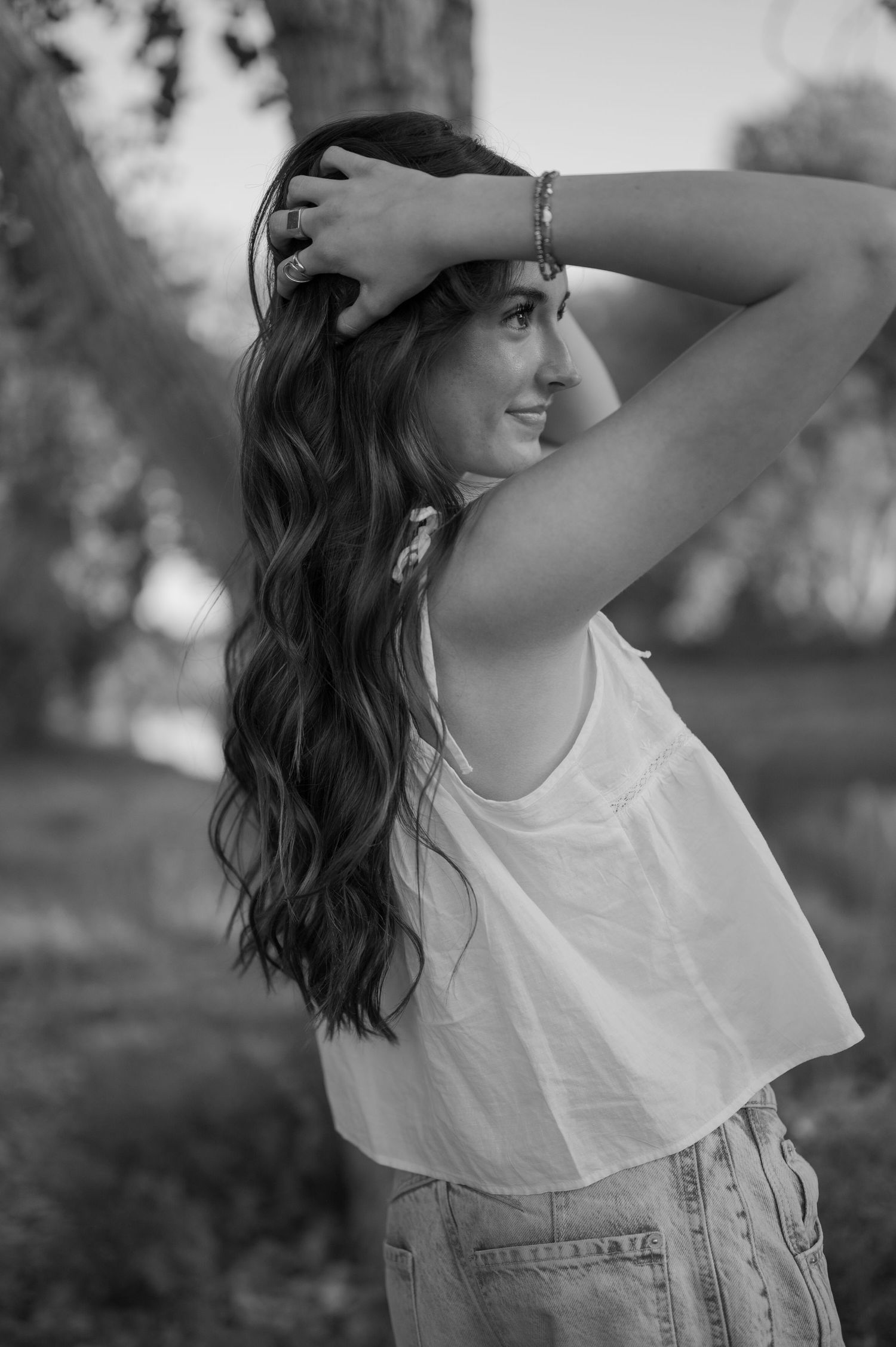 Black and white portrait of a high school senior with long wavy hair in a white crop top and jeans, smiling outdoors.