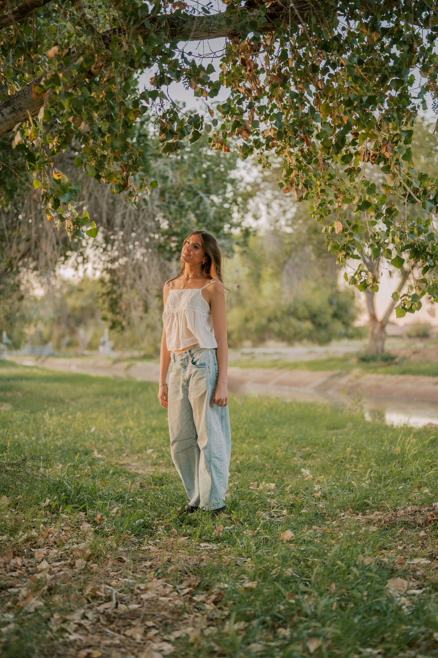 Senior portriars in white top and light jeans standing under tree canopy in sunlit park during golden hour.