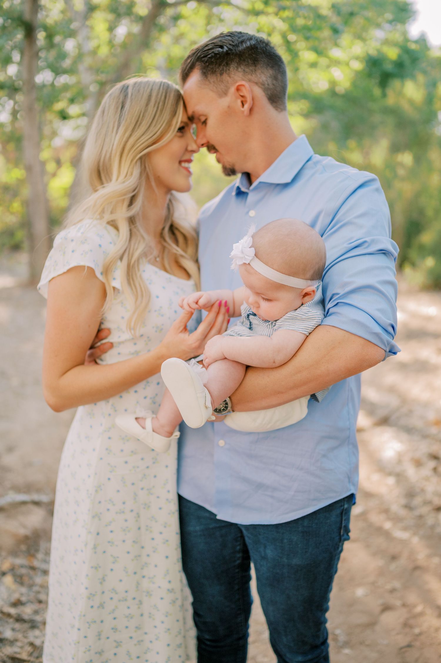A couple shares tender moments with their baby outdoors in a sun-dappled wooded setting. Phoenix Arizona family photographer.
