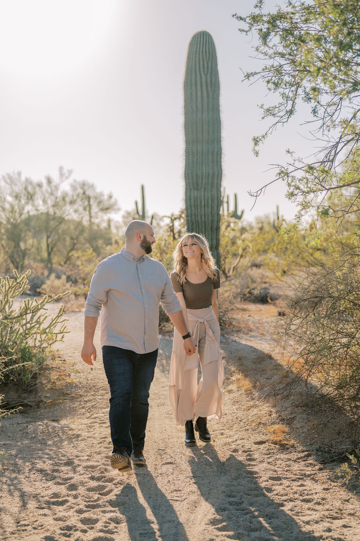 A couple walks hand in hand on a desert trail with tall saguaro cactus and desert vegetation in the background.