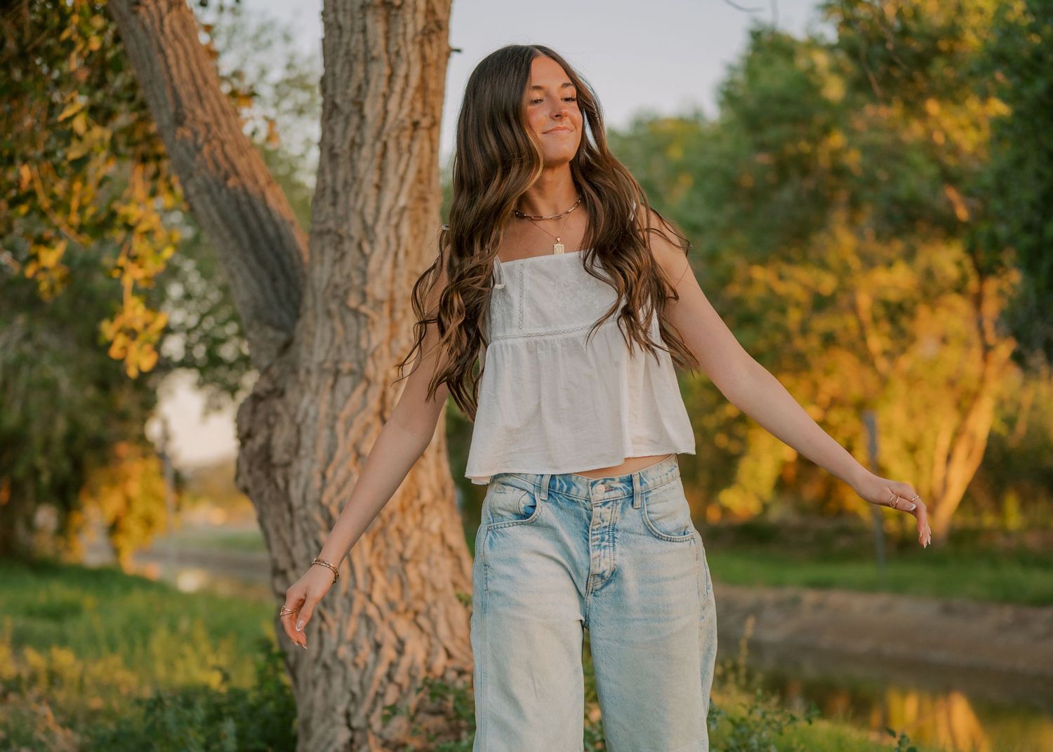 Senior girl with long wavy hair wearing a white crop top and light jeans smiling outdoors near trees.