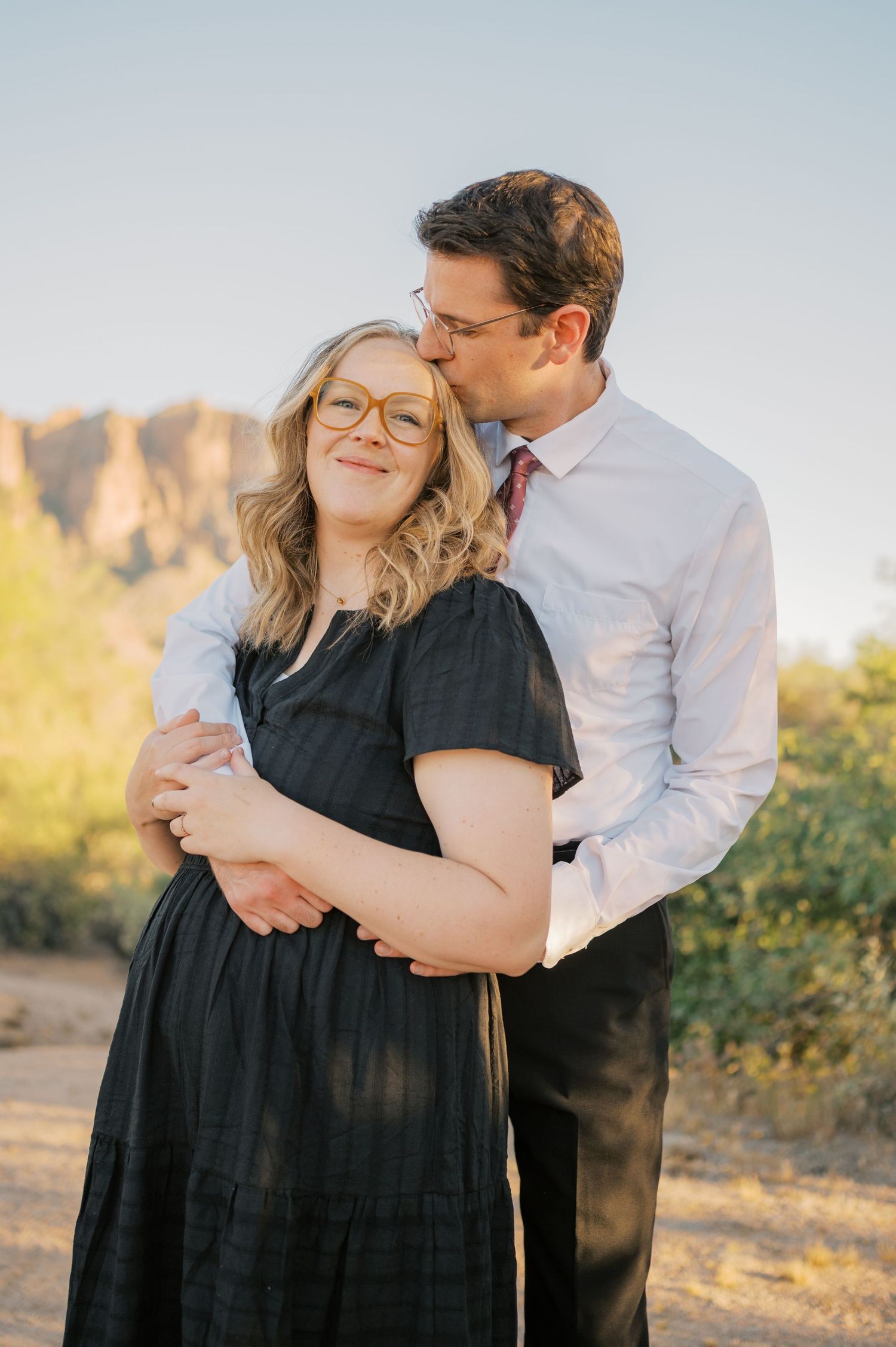A couple embraces outdoors, woman in black dress with glasses smiling as man kisses her temple, desert landscape background.