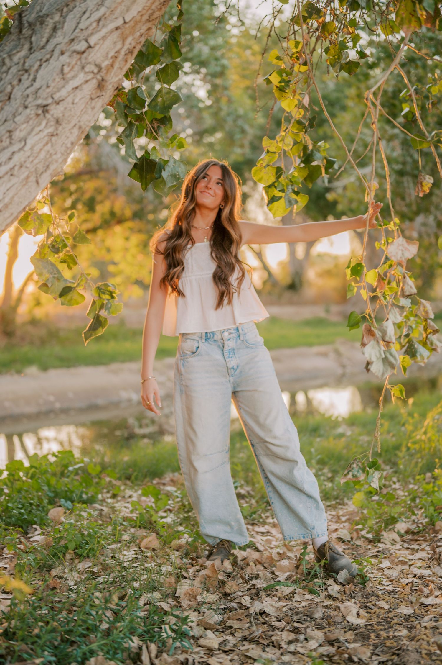Young woman in white crop top and wide-leg jeans poses under leafy tree branches in golden hour sunlight.