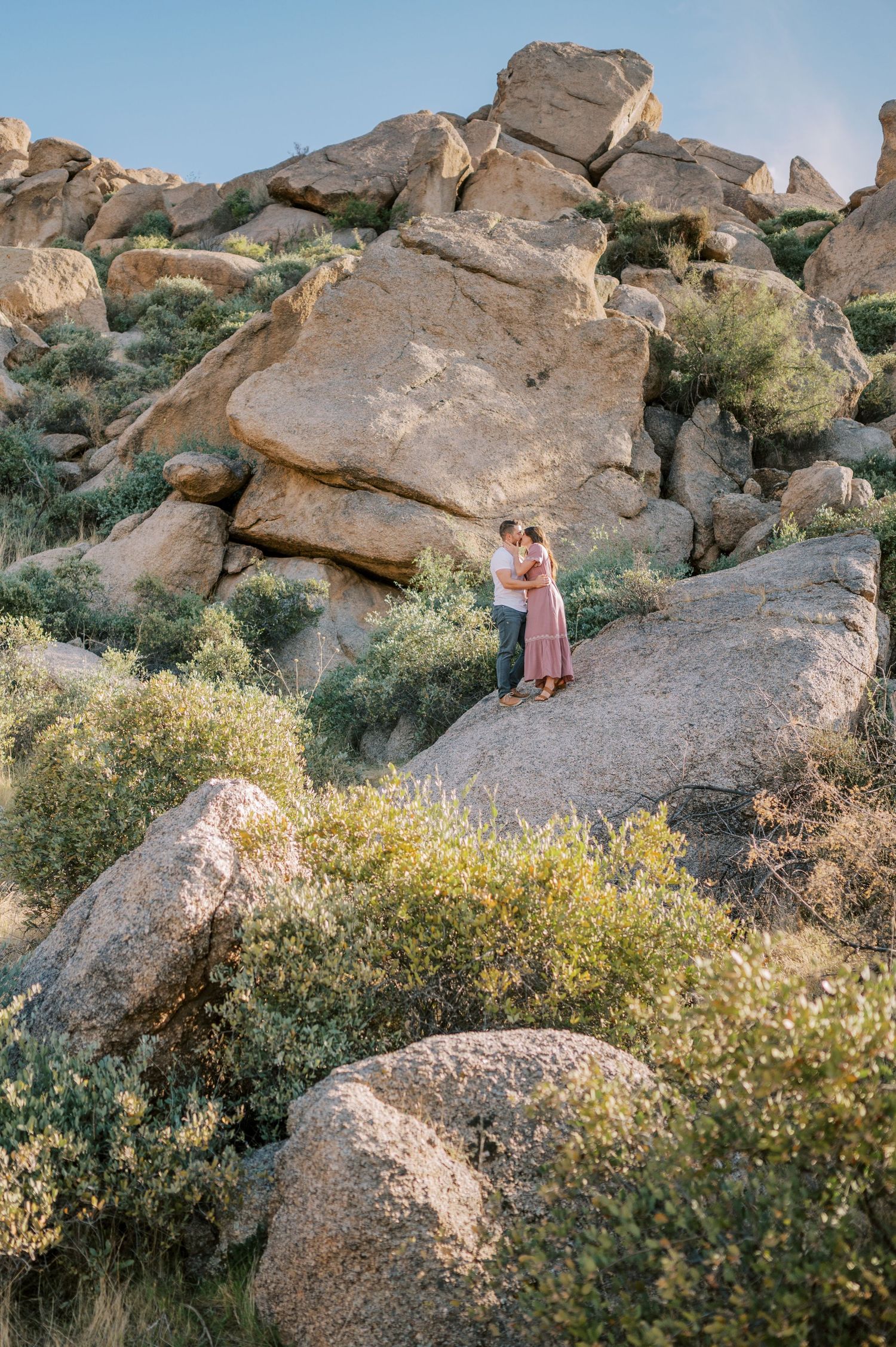 Couple embraces among large desert boulders and desert plants on a rocky hillside in golden sunset. Phoenix Arizona couples photographer.