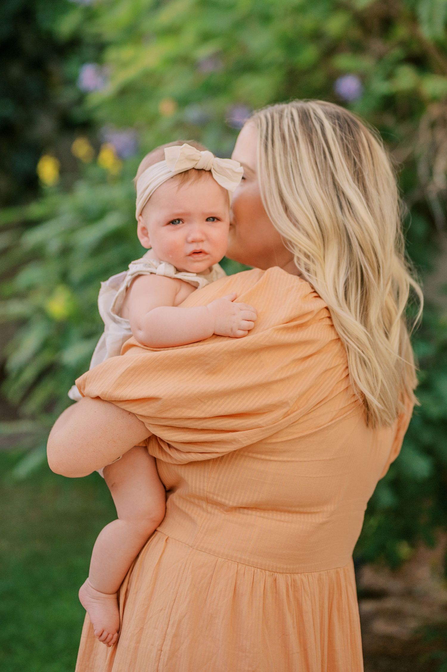 Parent in peach dress holding and embracing small baby outdoors in garden setting with green foliage background.