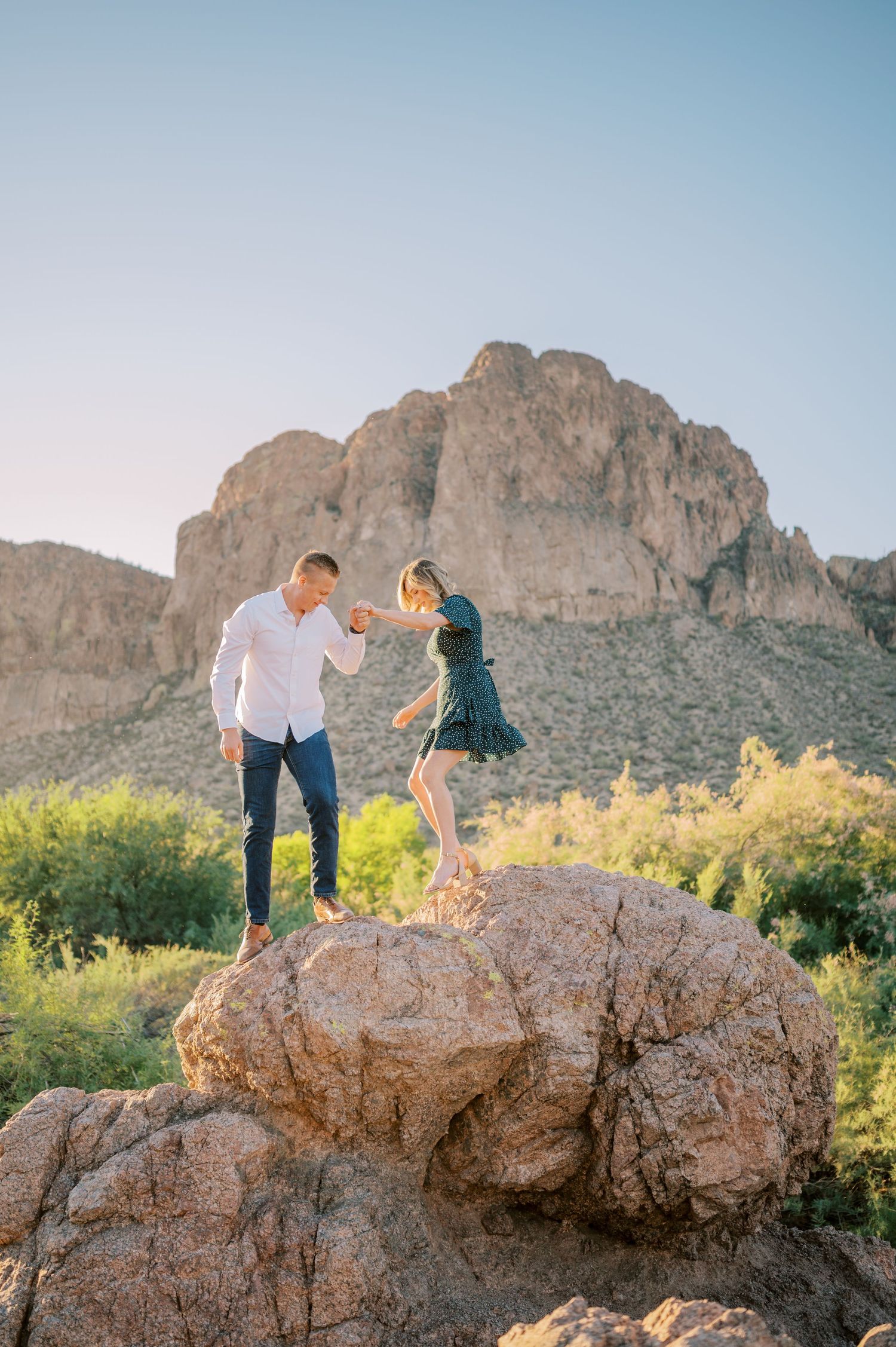 A couple explores rocky desert terrain with mountain cliffs in the background during golden hour. Phoenix Arizona couples photographer.