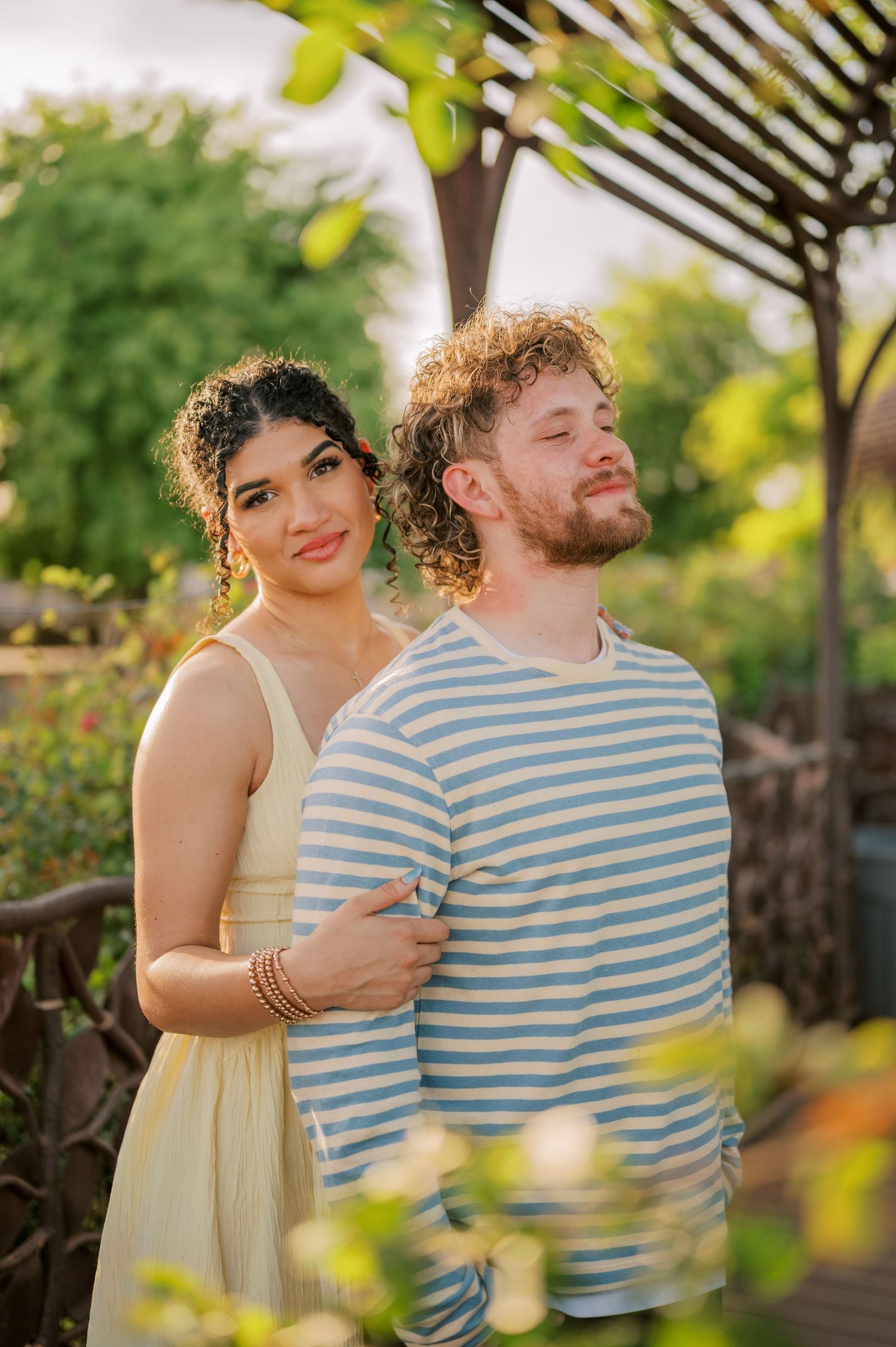 A couple poses outdoors in a lush garden, woman in yellow dress holds man in blue striped shirt as he tilts his head back.