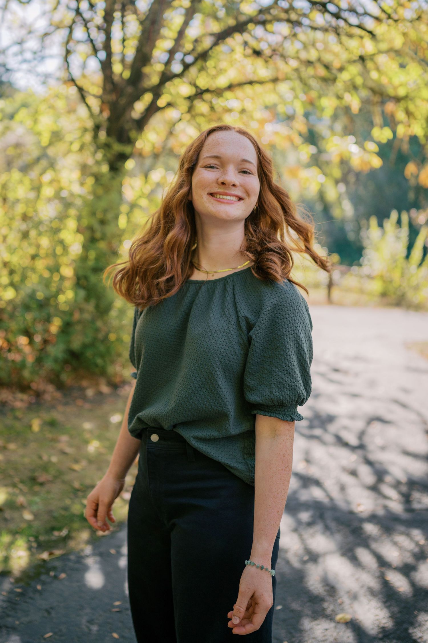 A person in a dark green top poses outdoors on an autumn day with golden sunlit trees in the background. Phoenix Arizona family photographer
