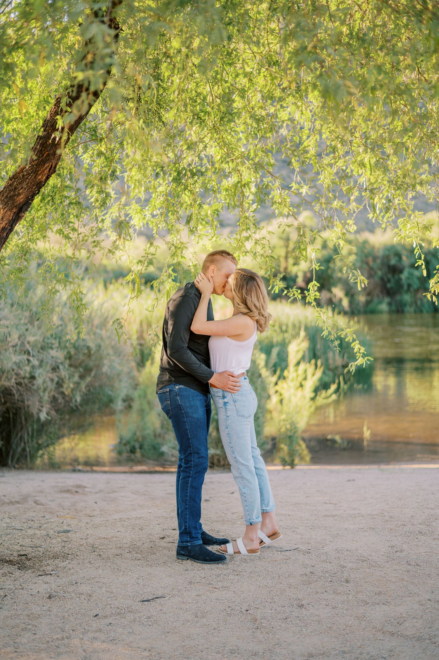 A couple shares a romantic moment by a riverside with overhanging willow trees in the background. Phoenix Arizona couples photographer.