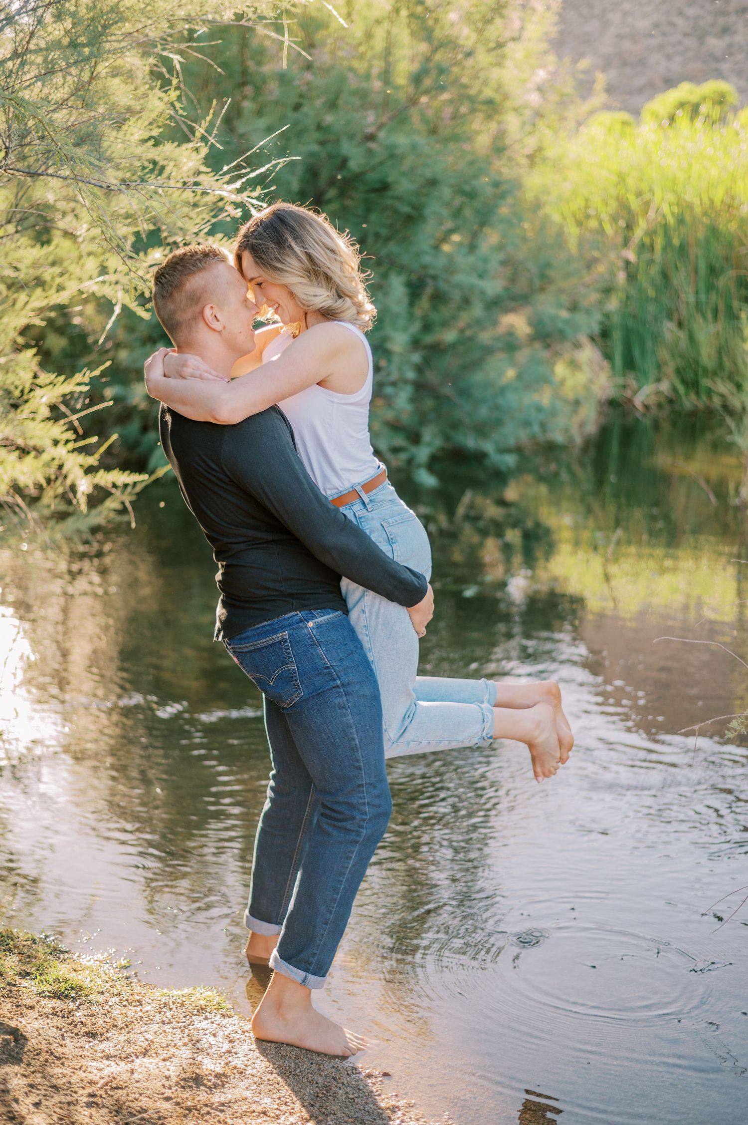 A couple embraces playfully by a serene pond in golden sunlight, creating a romantic summer moment. Phoenix Arizona couples photographer.