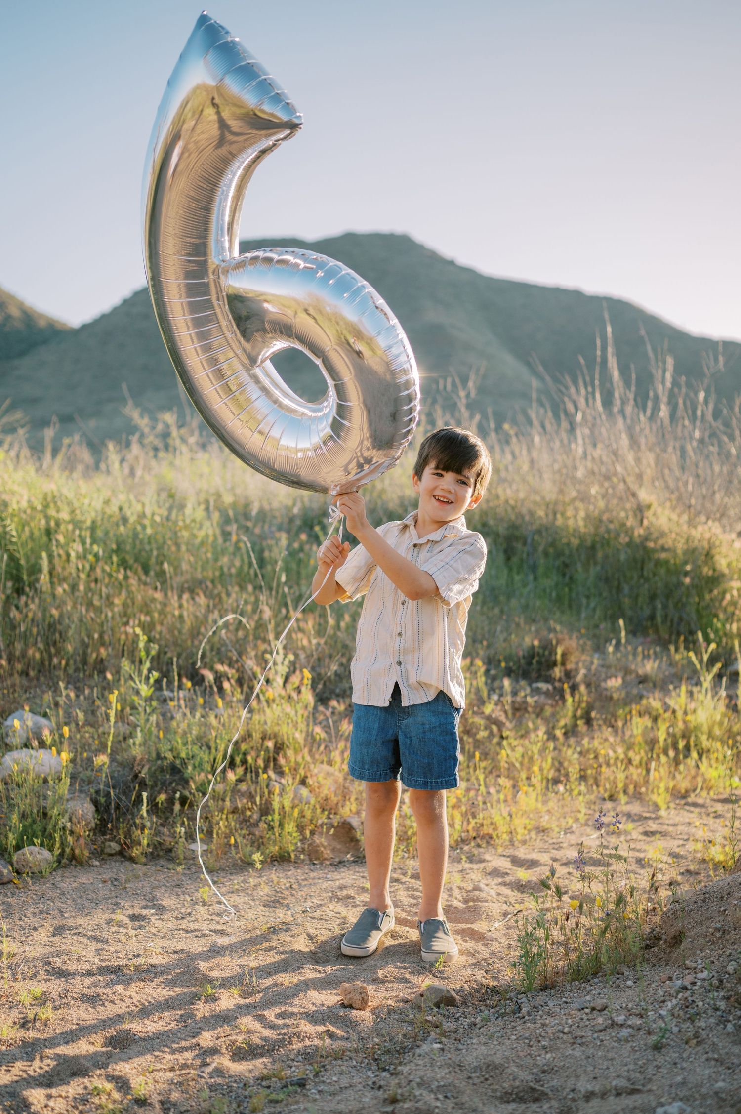 A large silver number 6 balloon is held outdoors against a mountainous landscape during golden hour. Phoenix Arizona portrait photographer.
