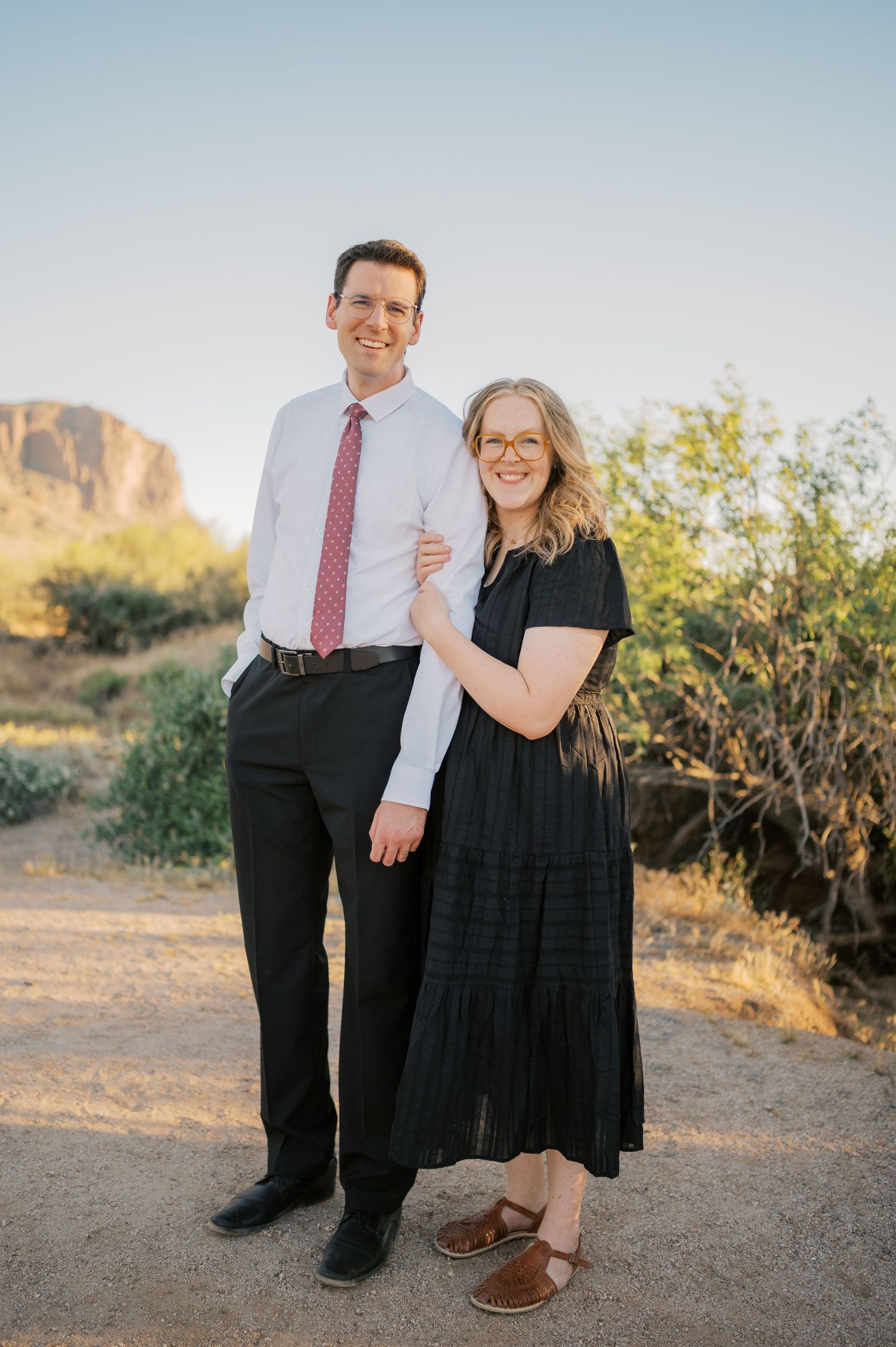 A smiling couple poses on a desert trail, man in dress shirt and tie, woman in black maxi dress, Arizona landscape behind.