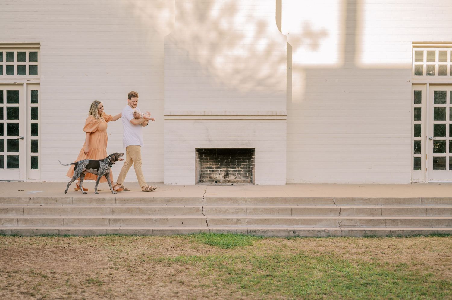 A couple walks their dog together along a white building with large windows and a fireplace. Phoenix Arizona family photograper.