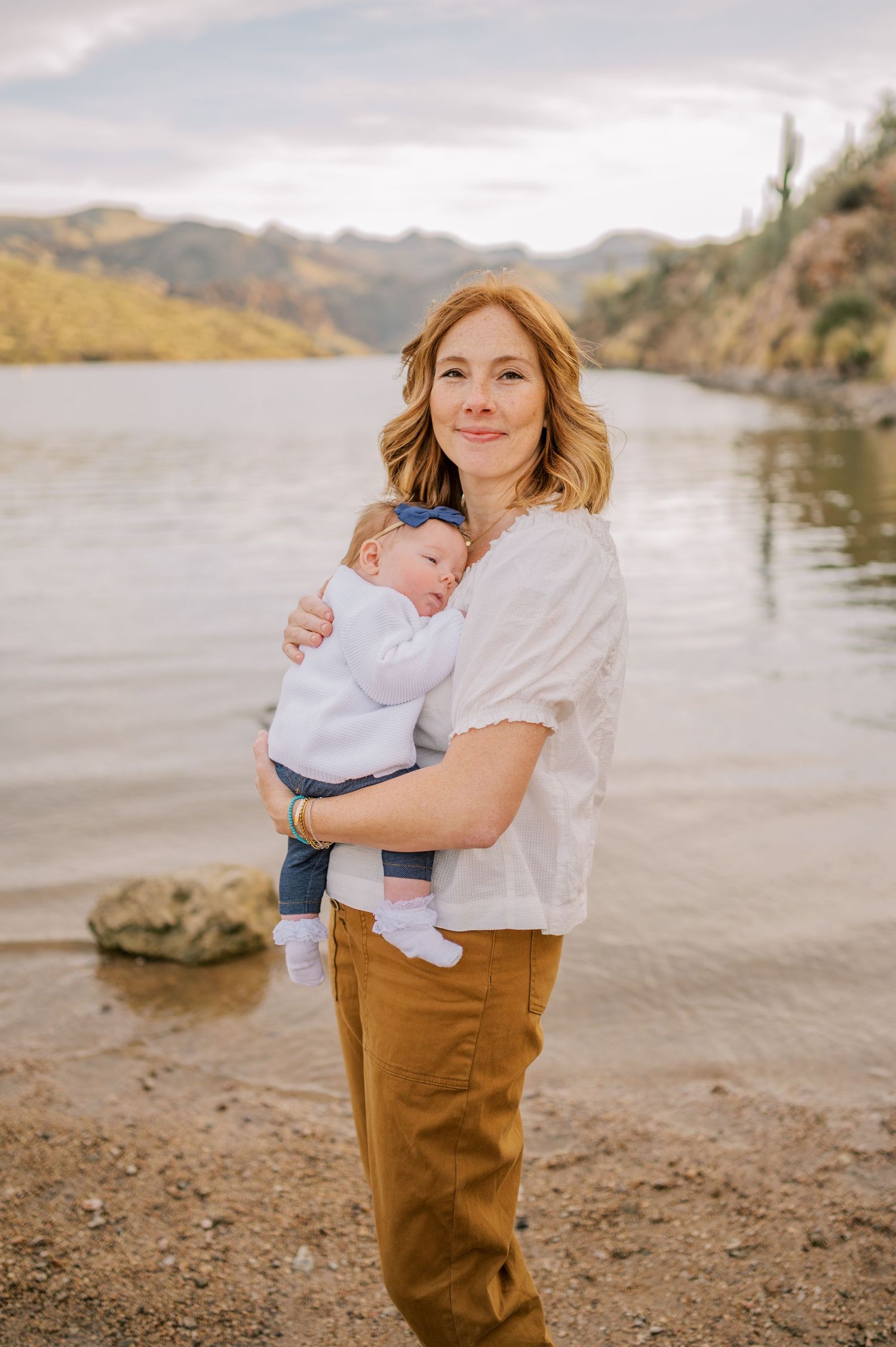 A parent and baby stand on the shore of a scenic lake surrounded by mountains on a cloudy day. Phoenix Arizona family photographer.