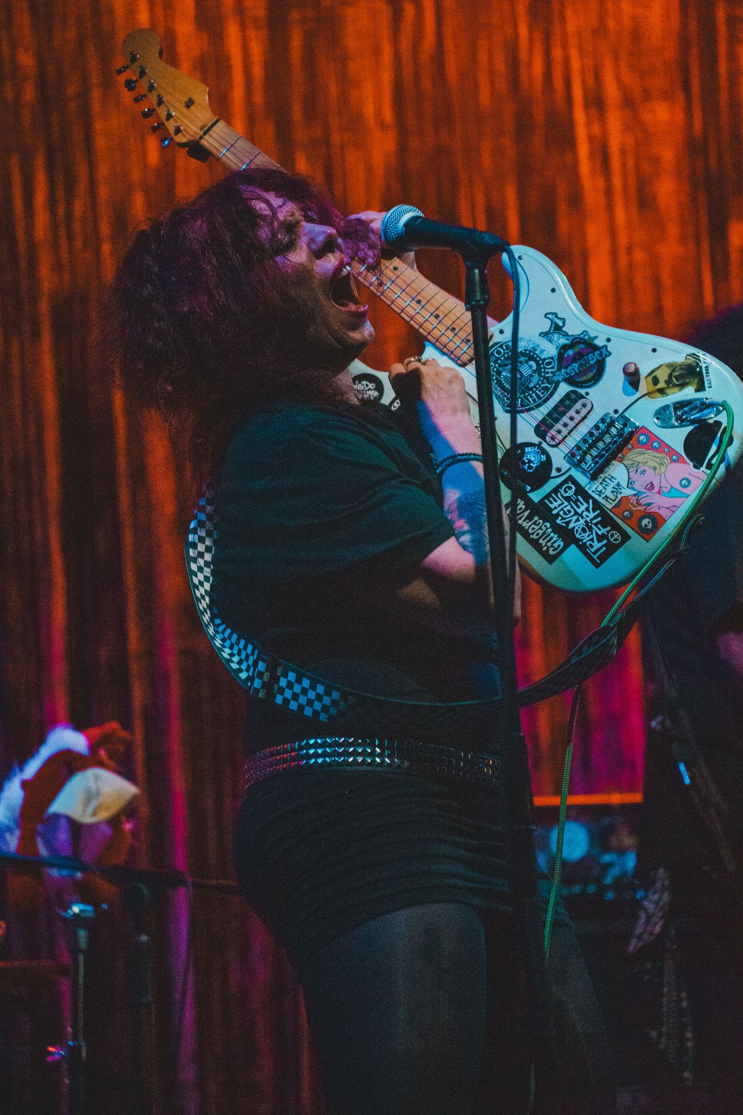 Rock musician singing passionately into microphone while holding a sticker-covered electric guitar on stage.