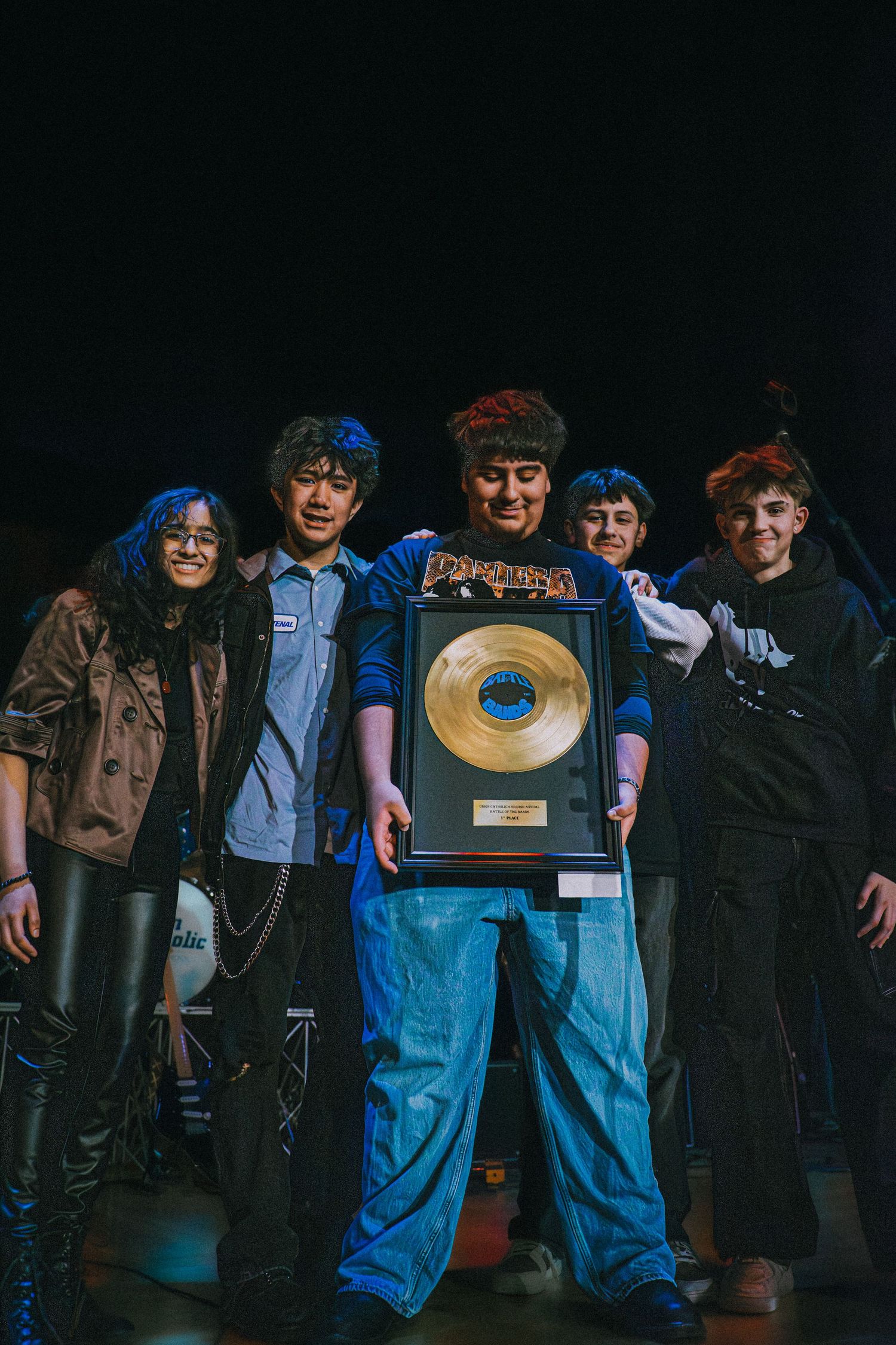 Group of young musicians posing on stage holding a gold record award in dark lighting.