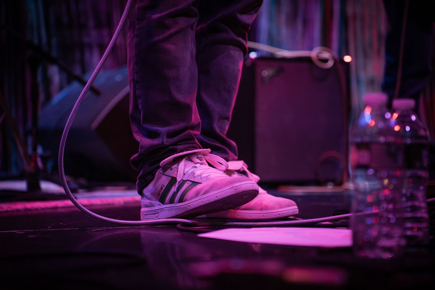 Close-up of white sneakers on a performer standing on a purple-lit stage with equipment nearby.