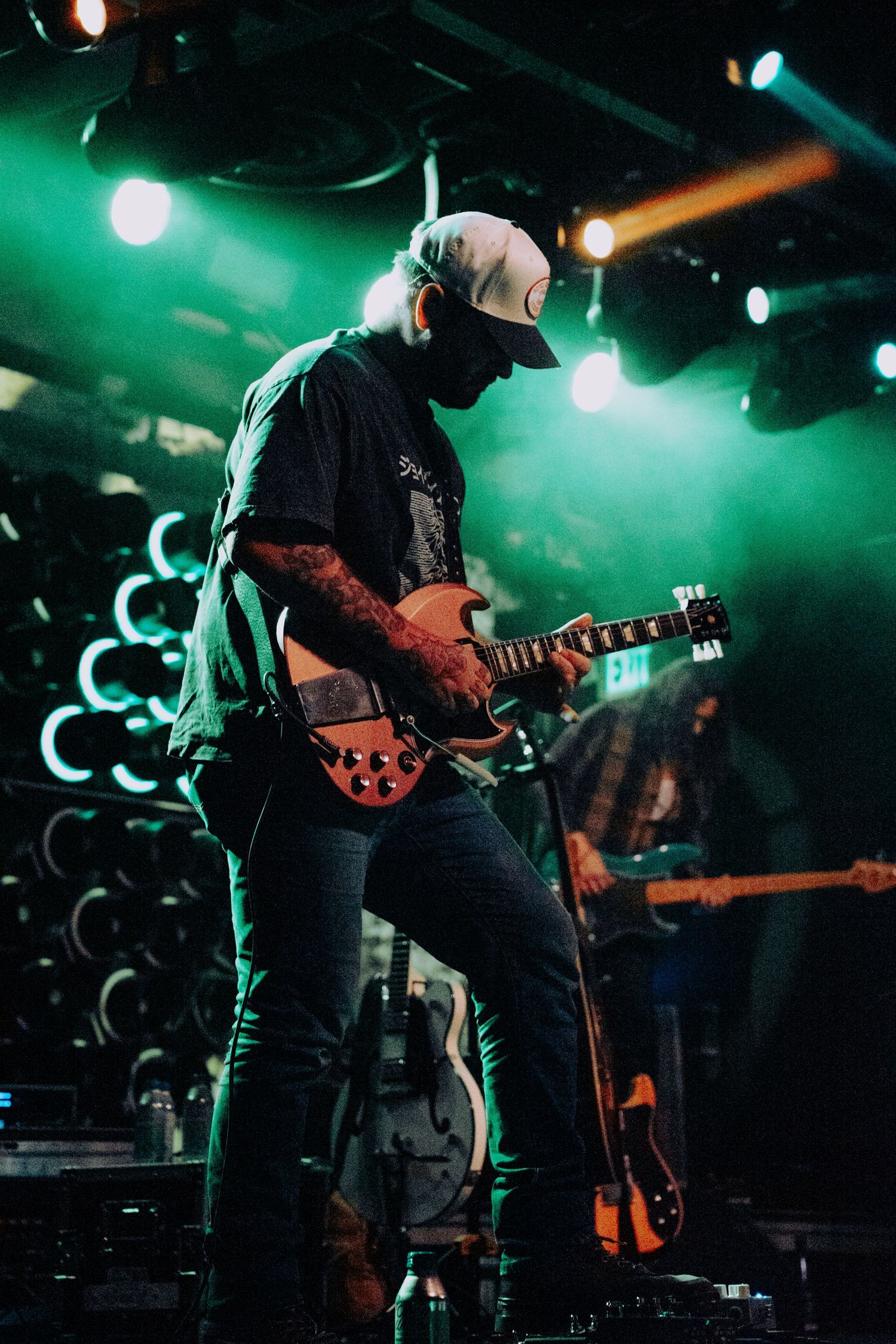 Guitarist performing on stage with green lighting, wearing black t-shirt and white cap at live concert.