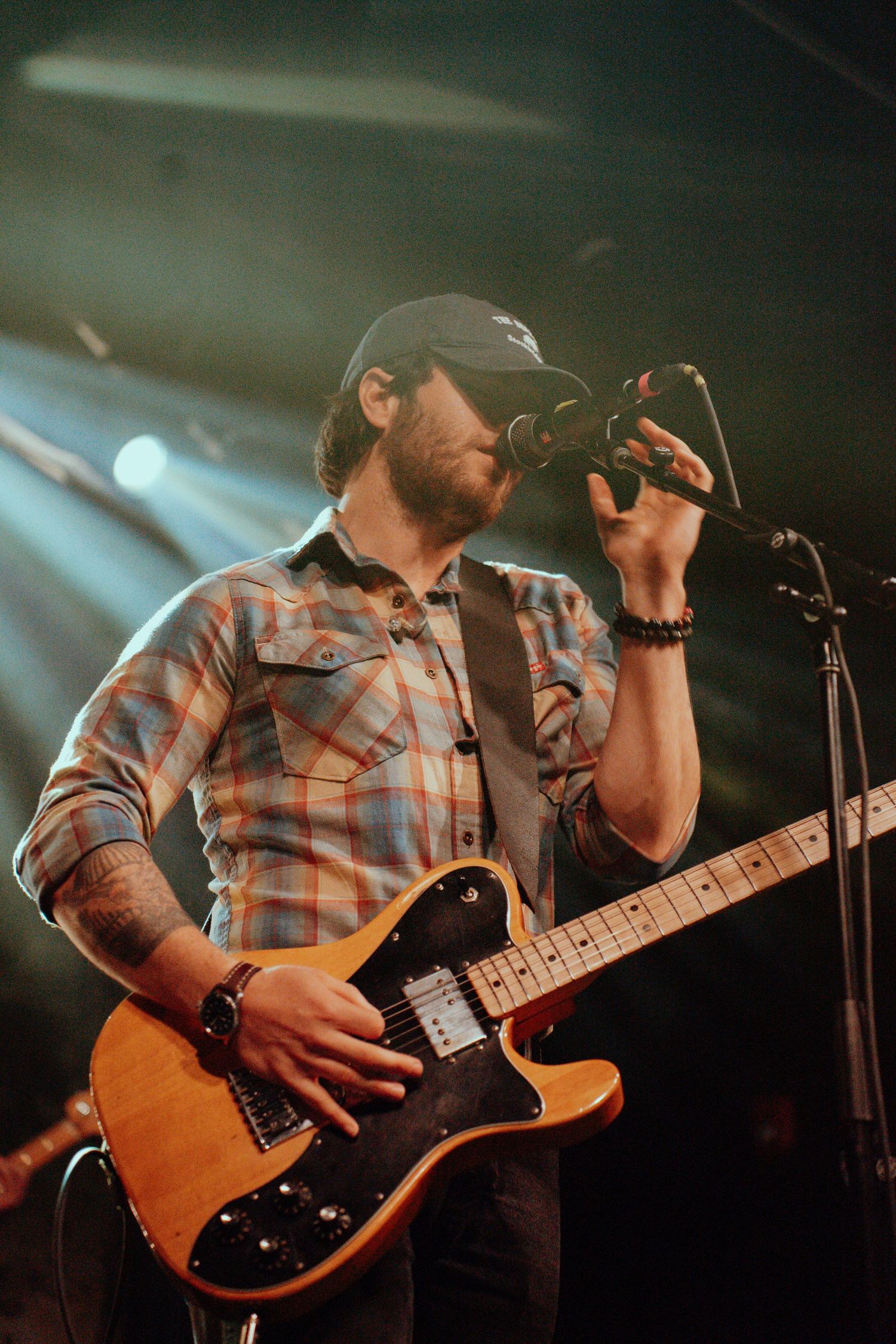 Bearded male guitarist in plaid shirt singing into microphone while holding a Telecaster guitar on stage.
