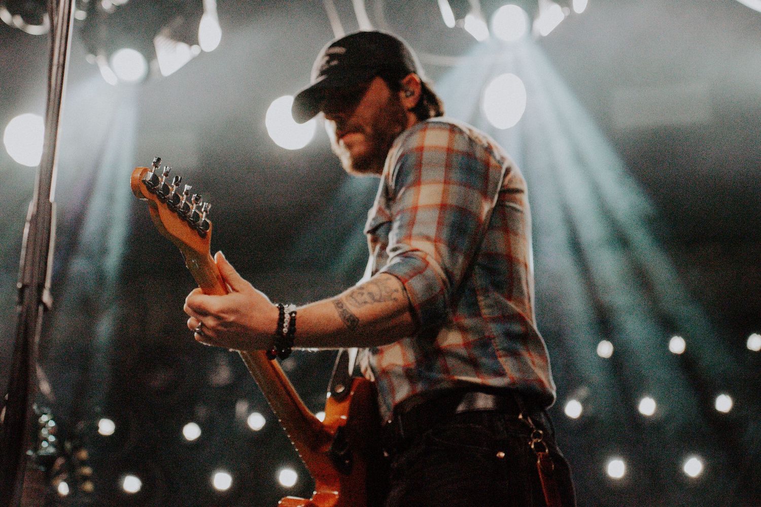 Guitarist performing on stage under dramatic spotlights wearing plaid shirt and cap.