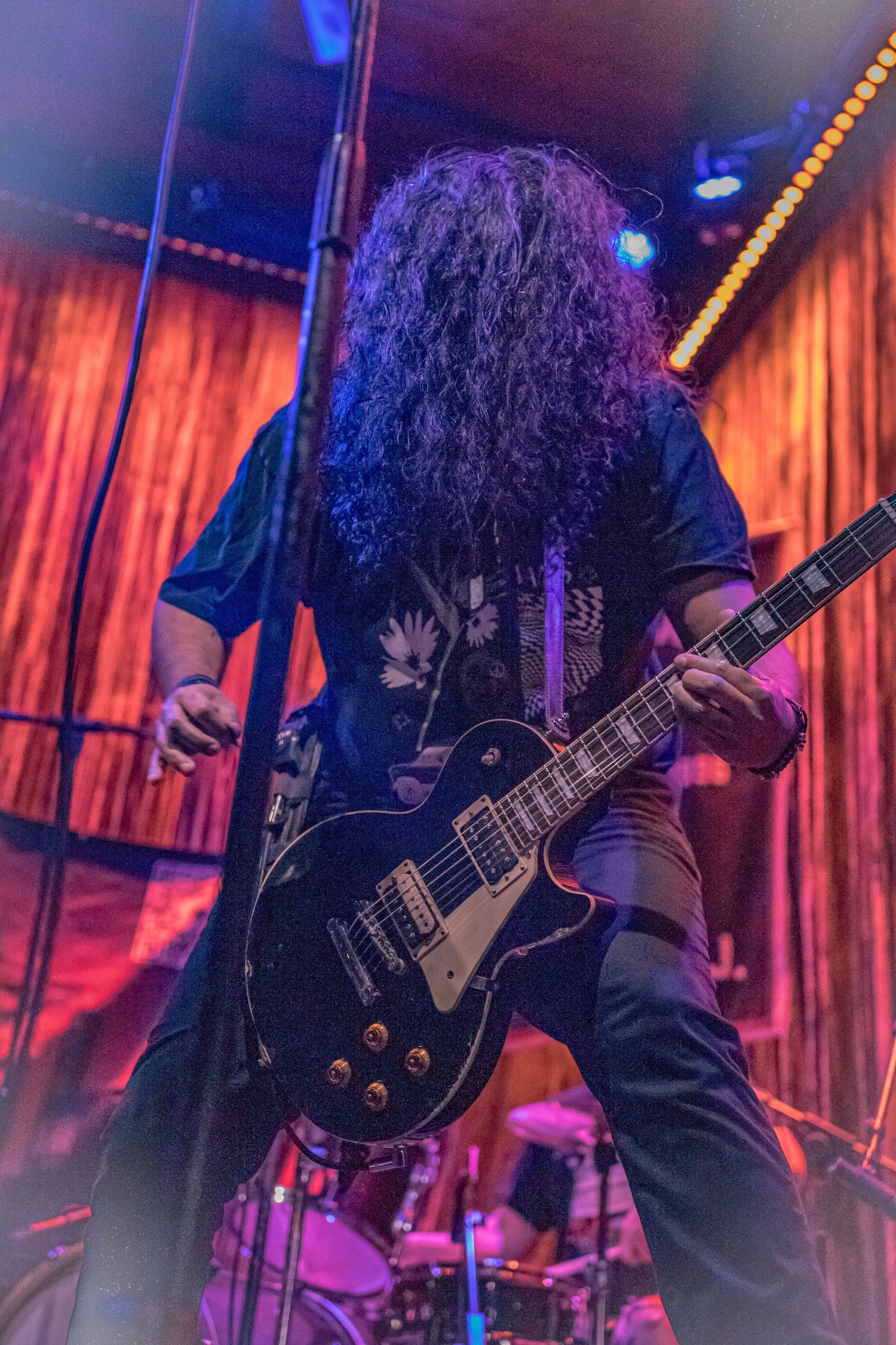Long-haired rock guitarist performing on stage with a Les Paul guitar under red and purple lights.
