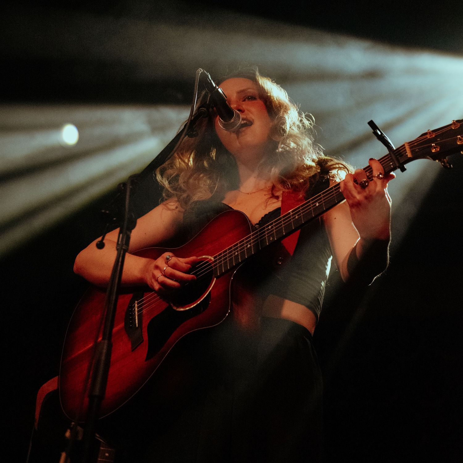 Female guitarist singing on stage, illuminated by dramatic spotlights while playing an acoustic guitar.