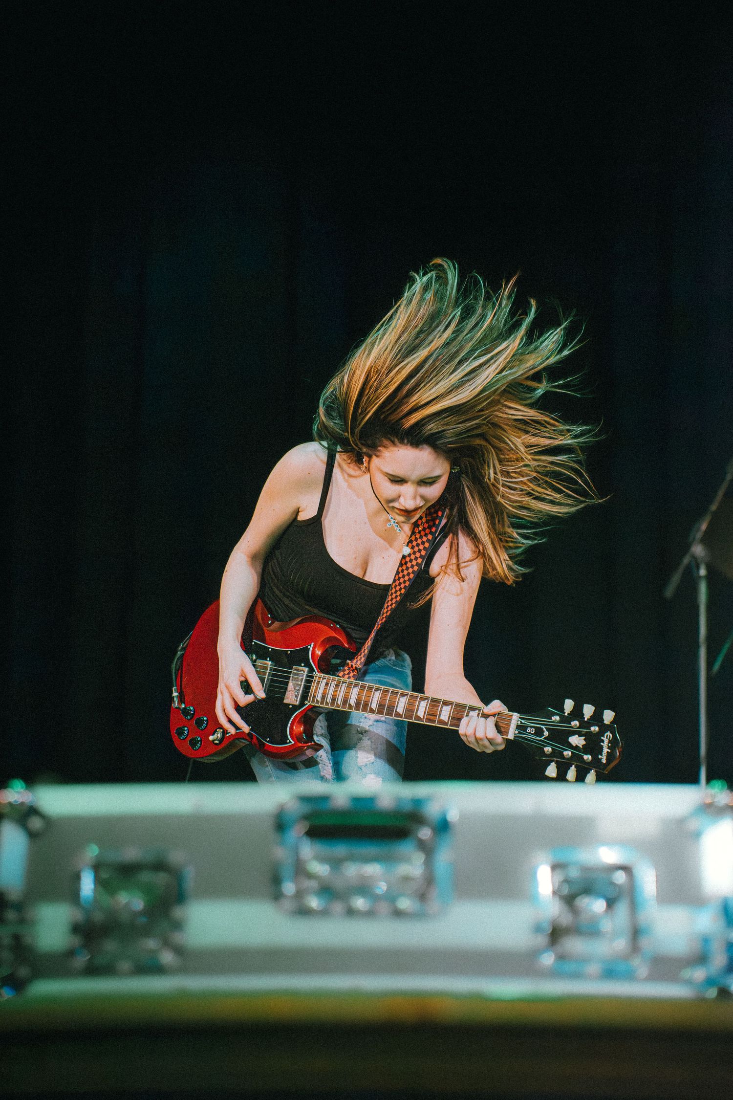 Female rock guitarist jumping on stage, hair flying, playing a red electric guitar energetically.