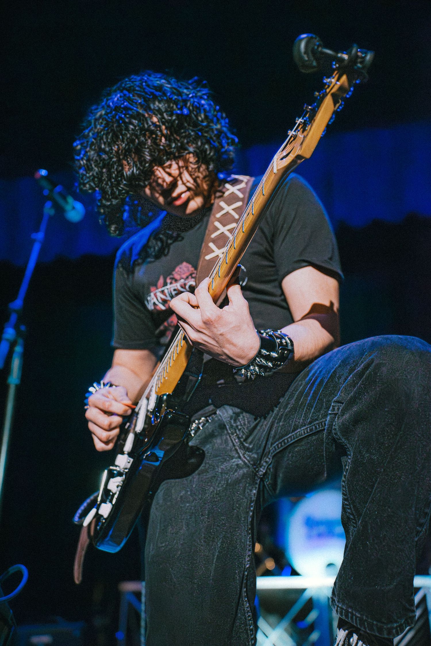 Curly-haired guitarist performing on stage, intensely playing an electric guitar under blue stage lights.