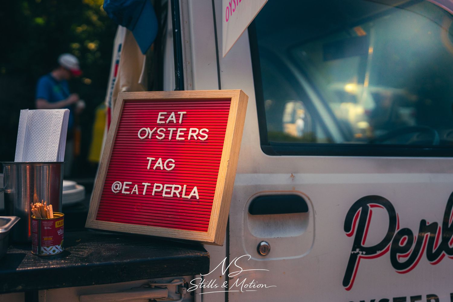 Red letter board sign reading Eat Oysters Tag @EatPerla displayed beside a food truck at night.