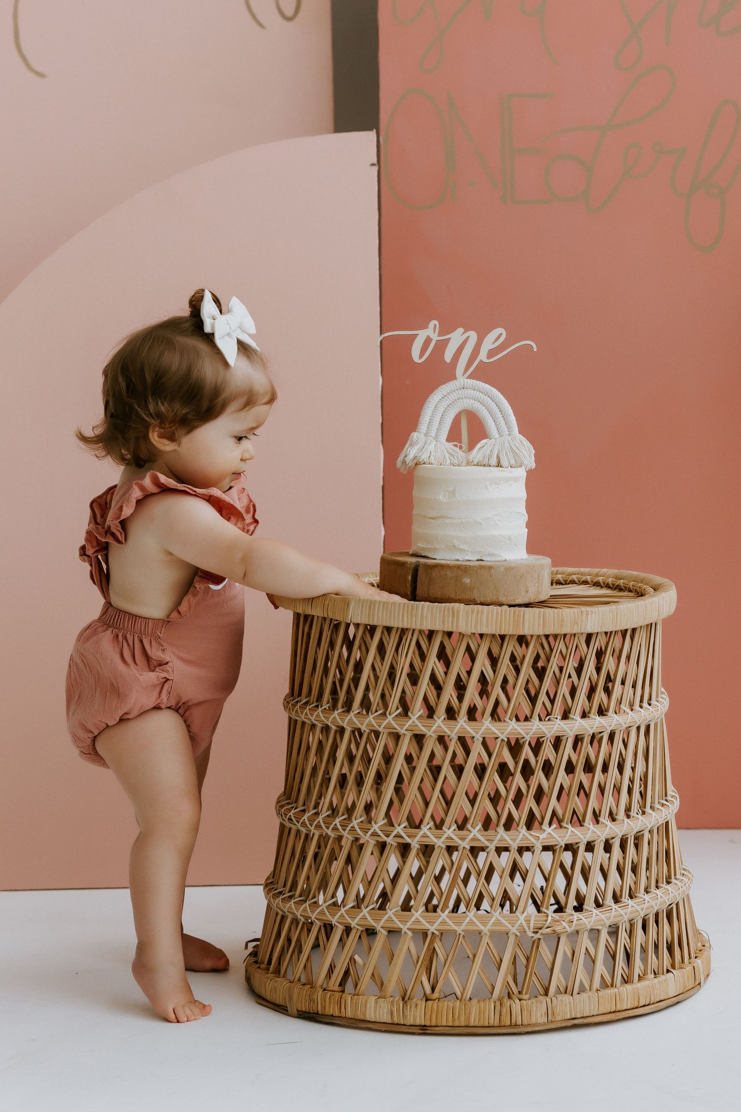 Baby exploring birthday cake setup with woven rattan base and pink backdrop.