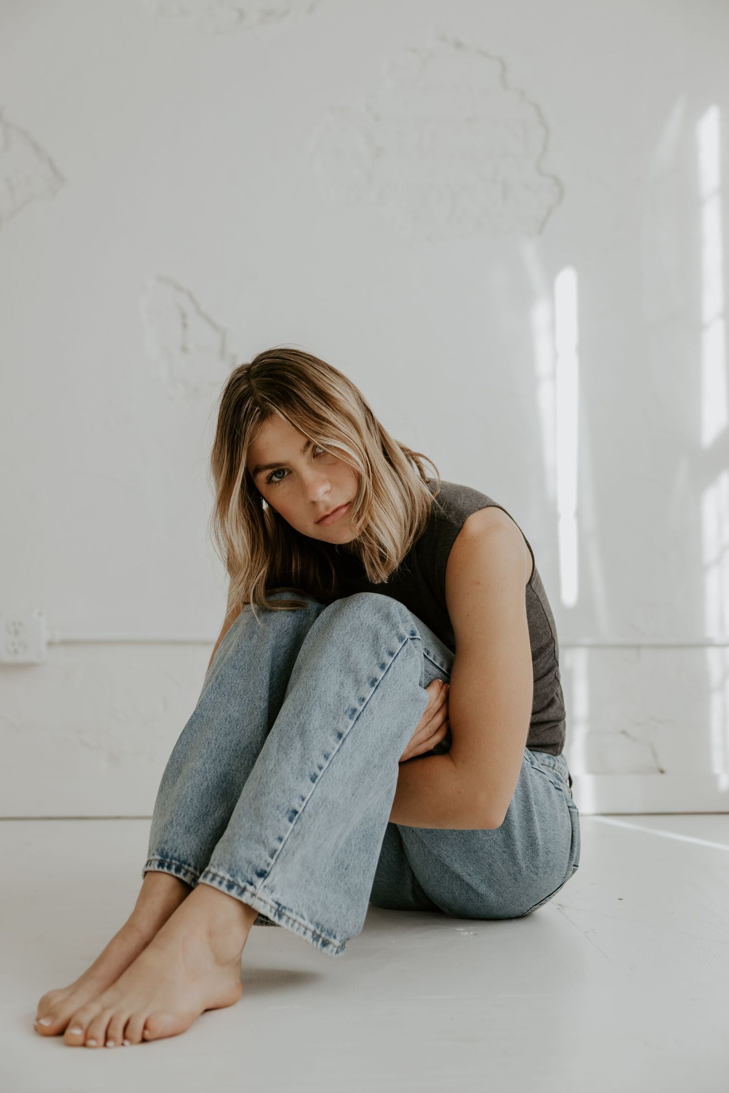 Person in casual attire sitting on white floor wearing jeans and dark top in minimalist setting.