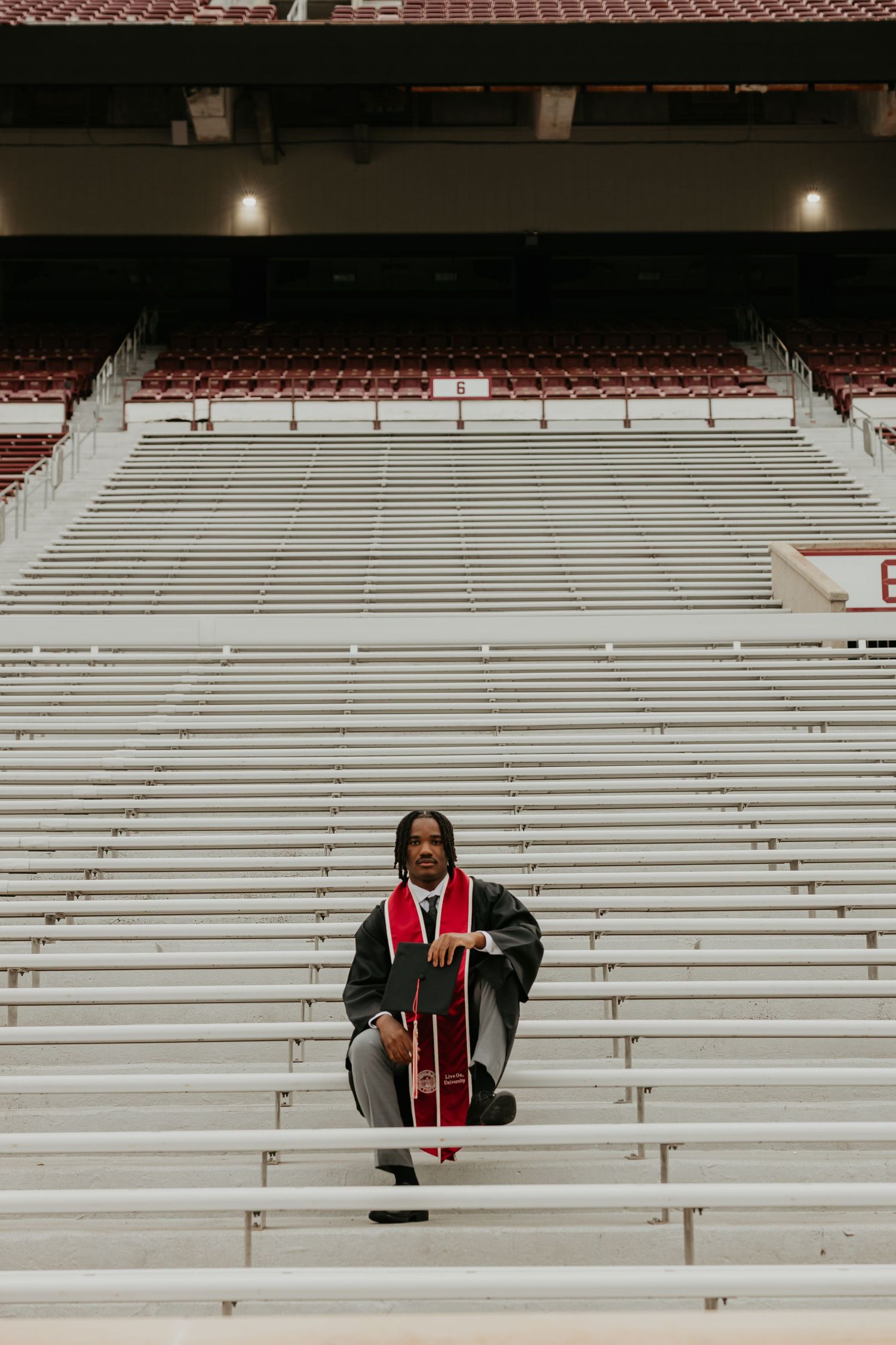 Graduate in red and black regalia sitting alone on stadium bleachers.
