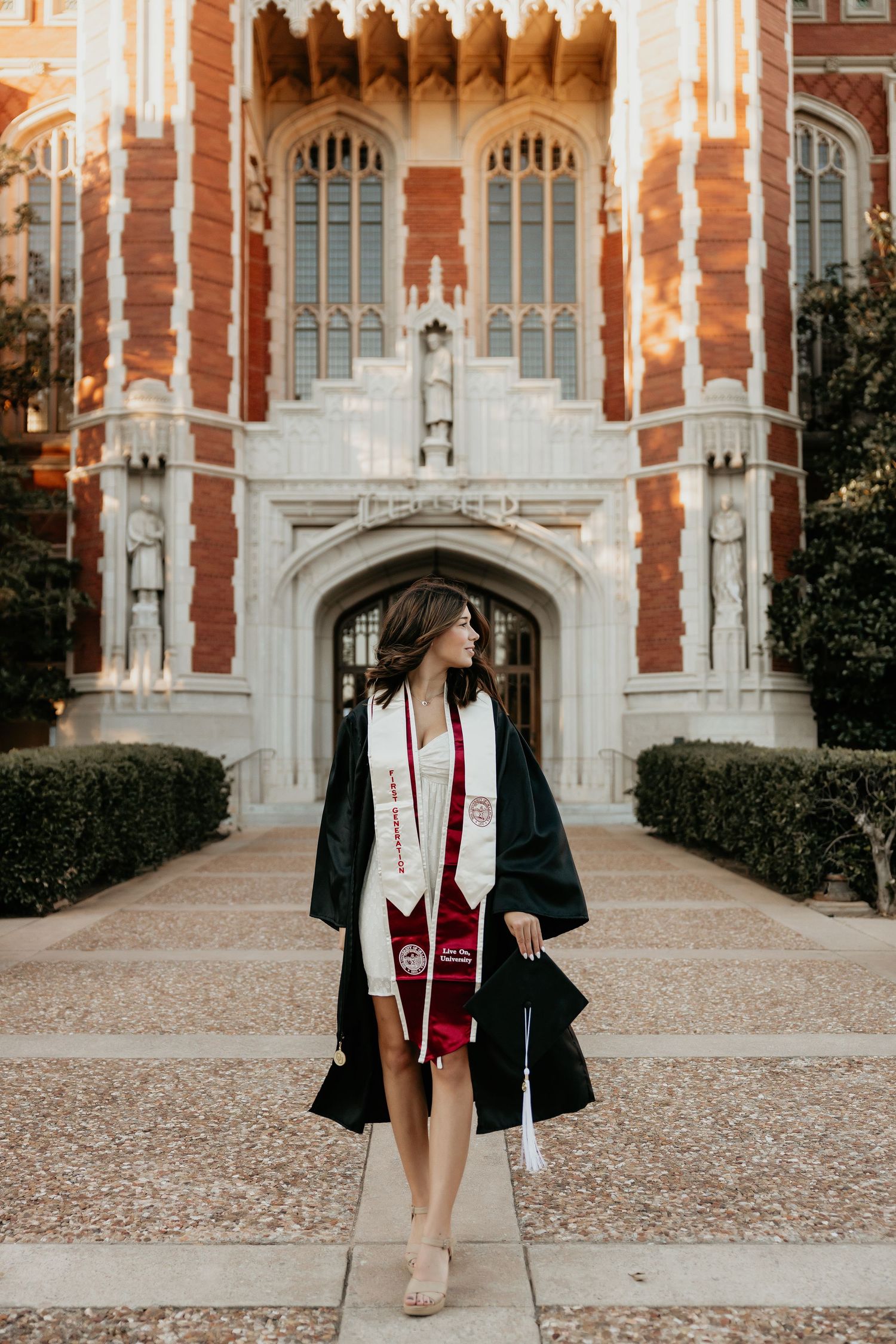 Graduate poses in cap and gown in front of ornate Gothic university architecture.