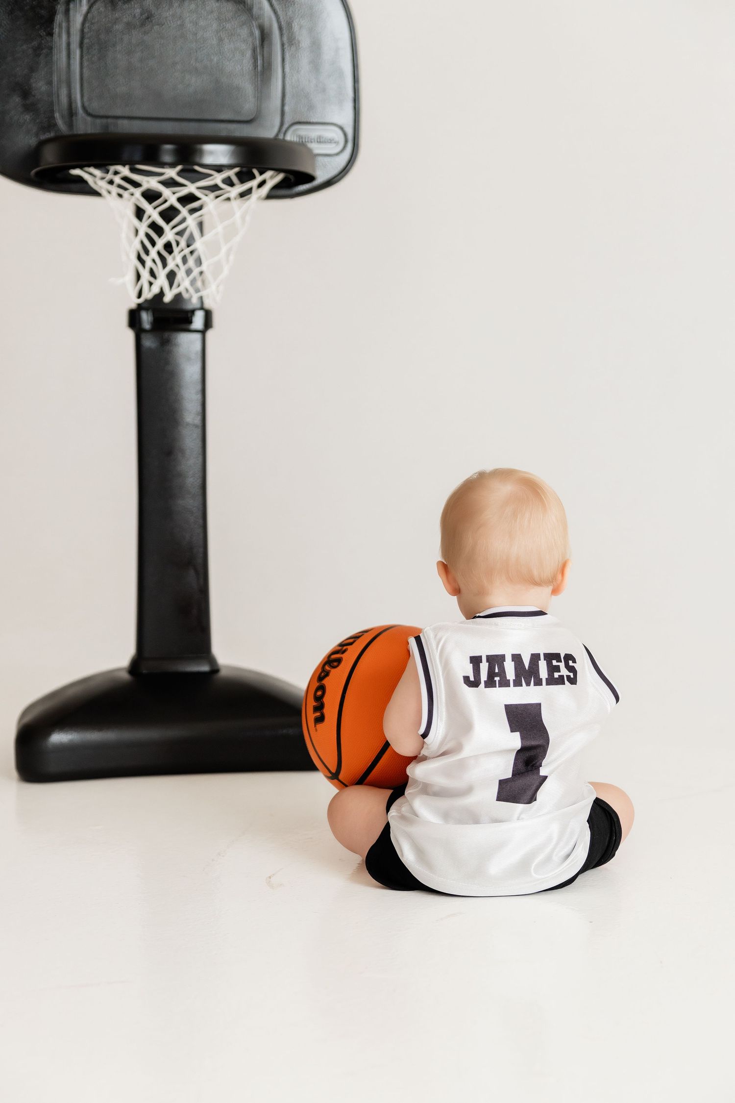 Basketball themed baby photo series with white jersey and basketball hoop against white background.