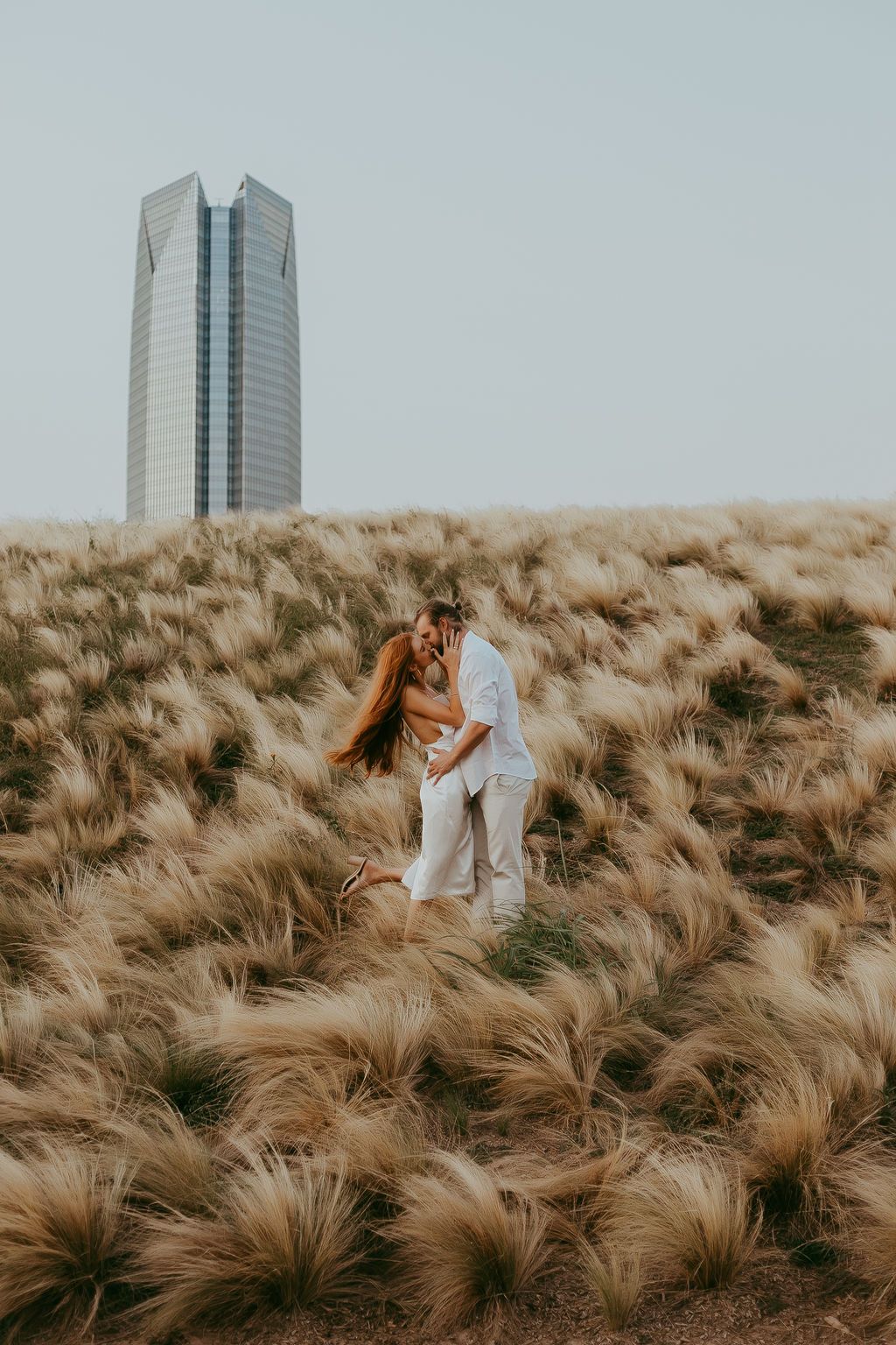 A couple embraces on a hill covered in tall grass with city buildings visible in the background.