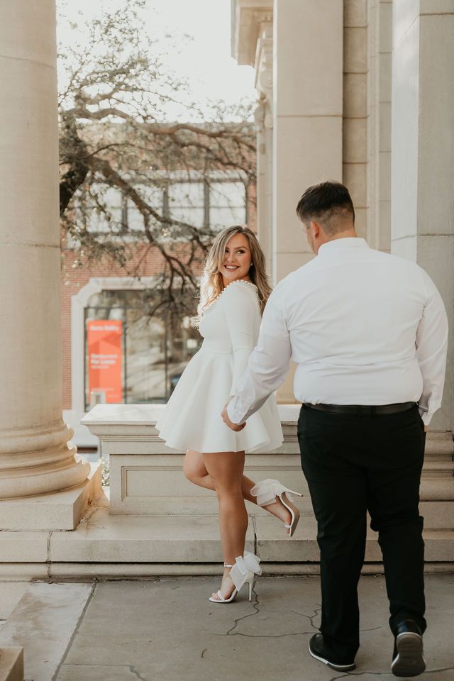 Playful engagement photo of couple posing by classical columns at sunset.