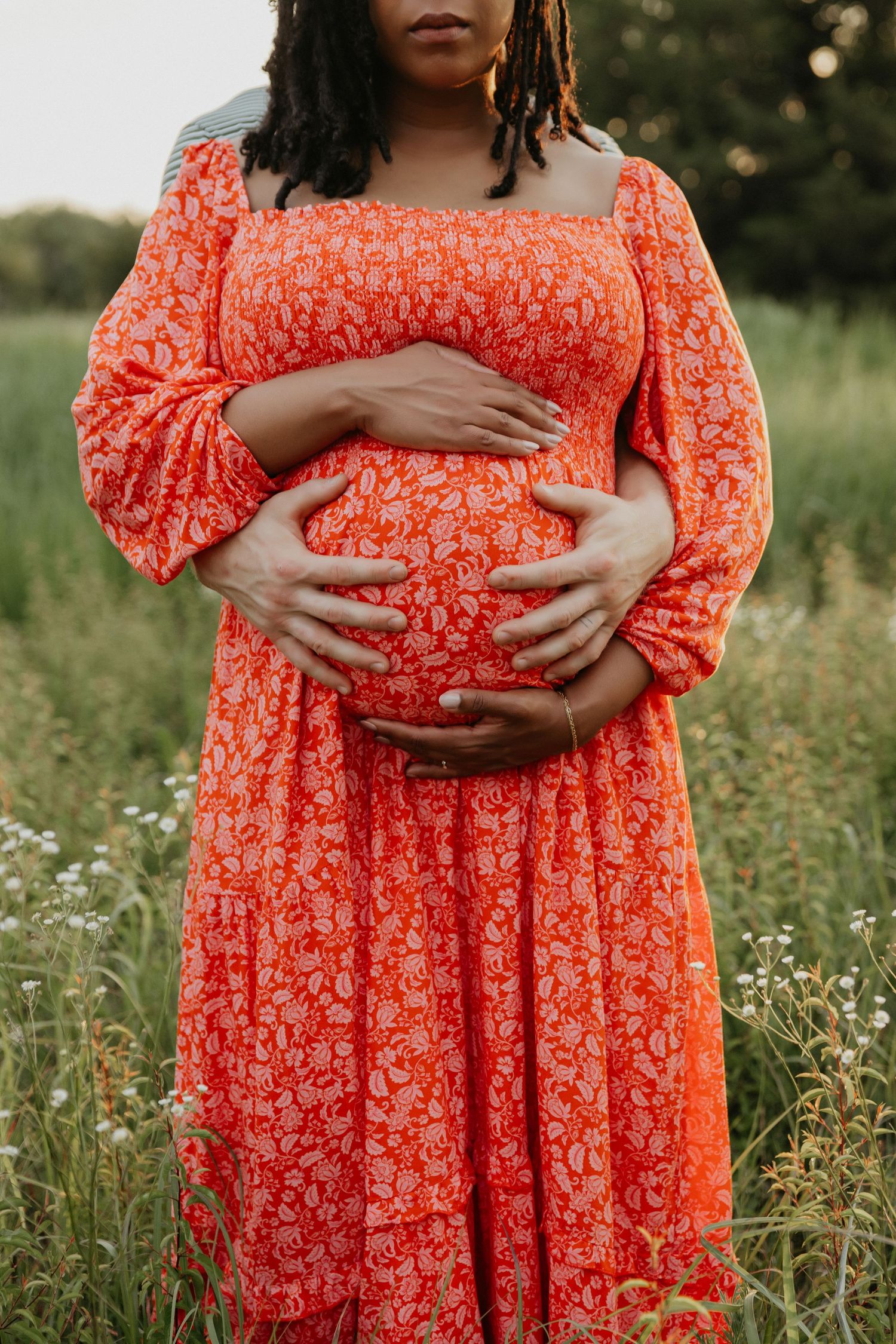 A person in a vibrant coral floral dress cradles their pregnant belly in a wildflower field.