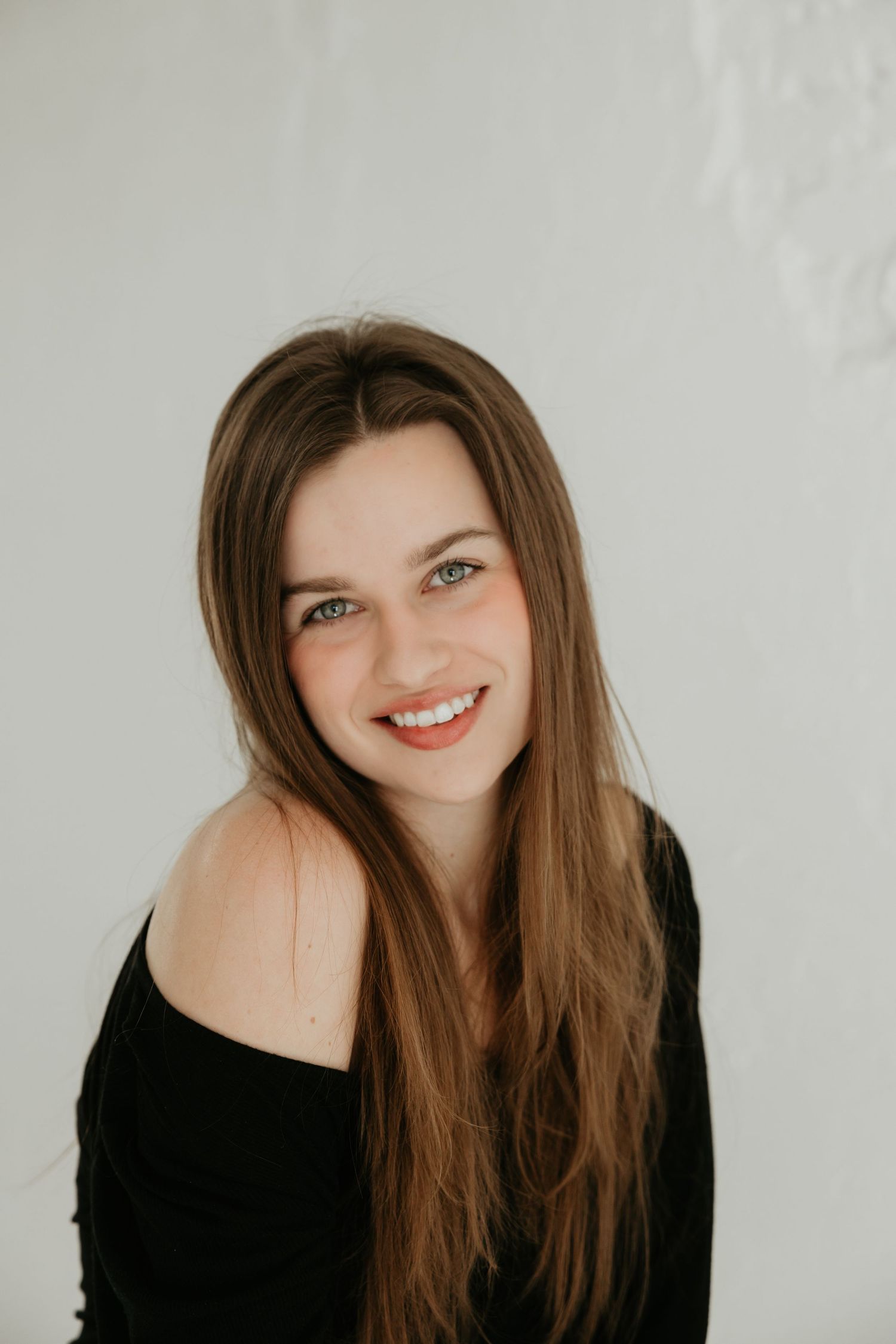 Portrait against white wall showing warm smile and long brown hair in black off-shoulder top.