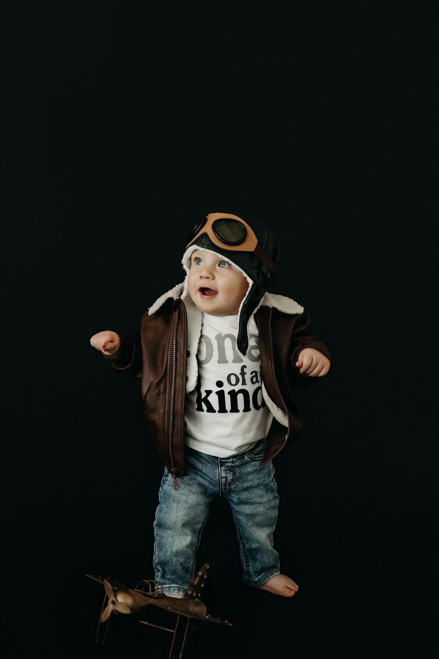 Baby in a brown jacket and jeans strikes a cool pose on black background.