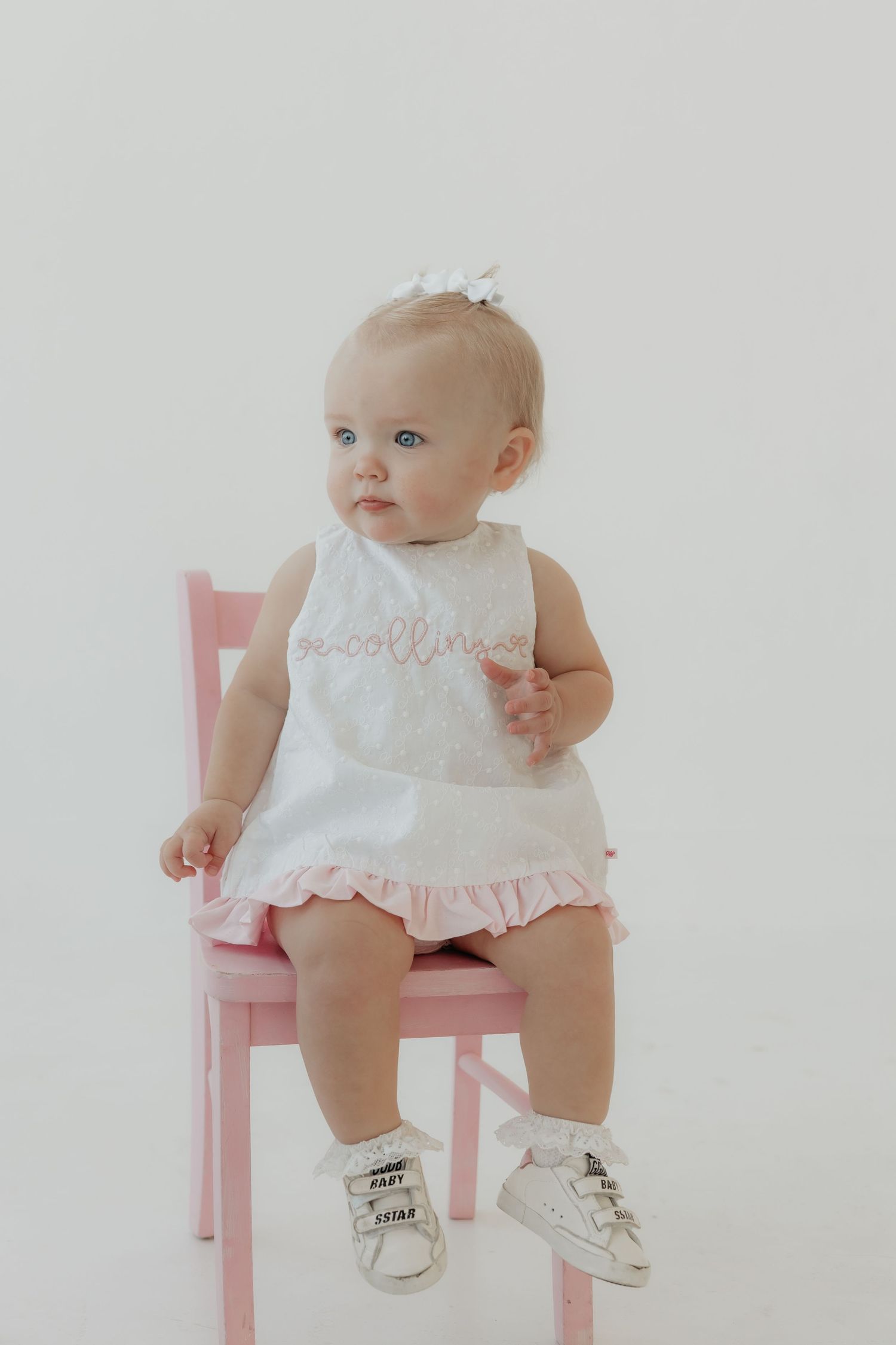 Baby in white dress sits on pink chair wearing gold shoes.