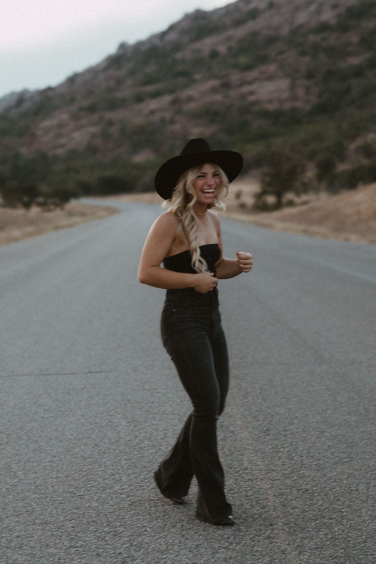 Person in dark clothing dances playfully on an empty mountain road at sunset.