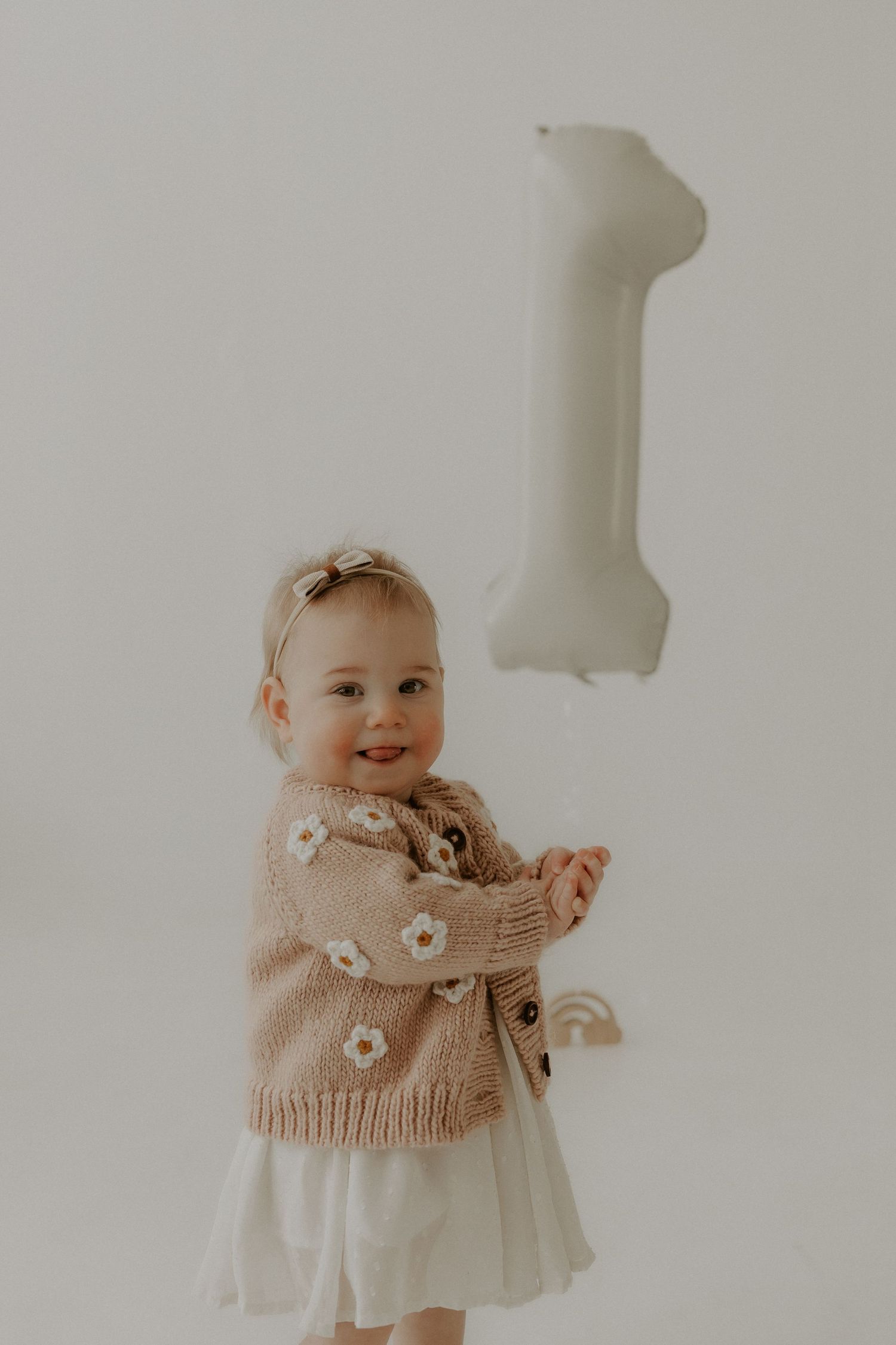 Baby wearing white dress and knit sweater with number 1 balloon.